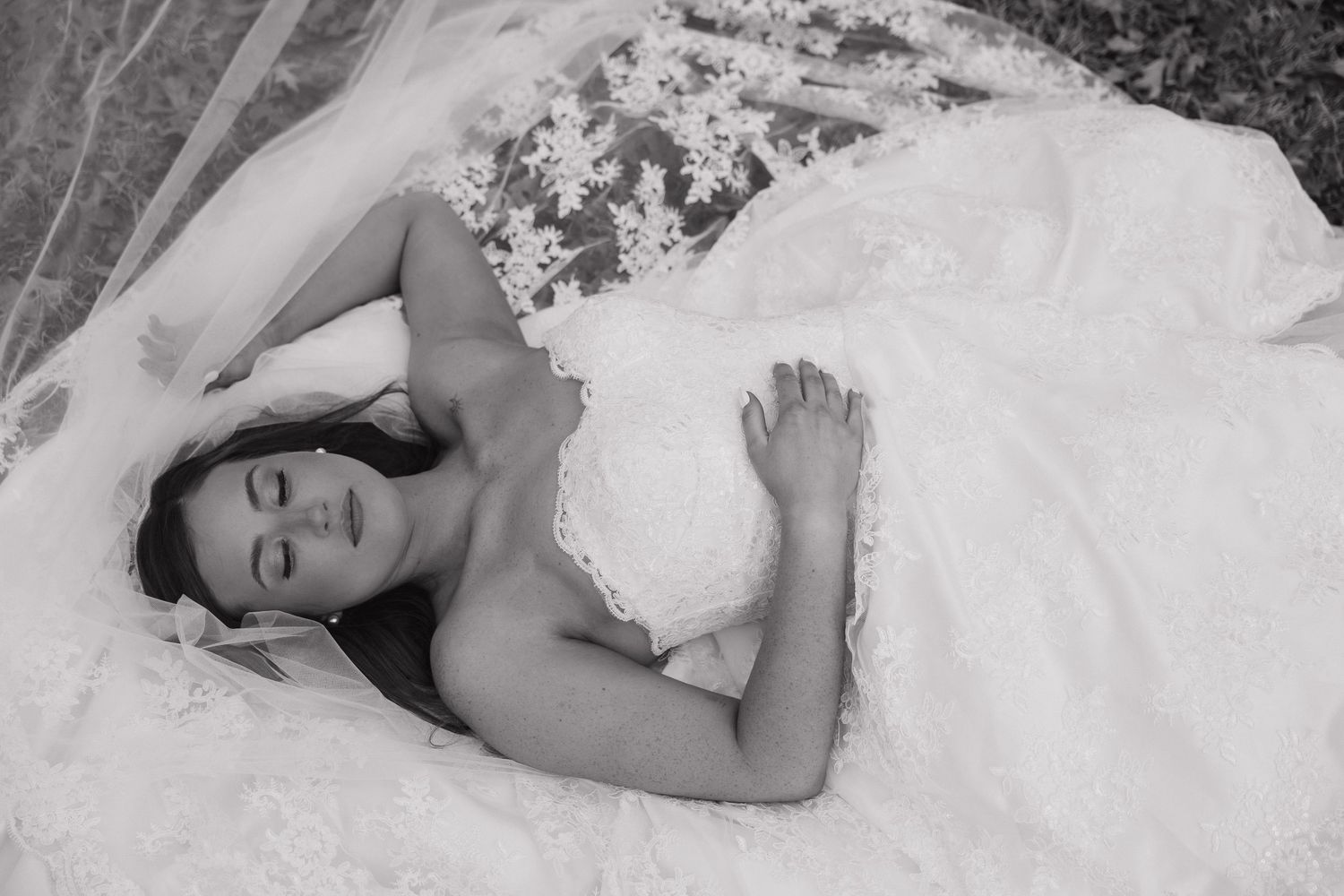 A bride in a white lace wedding dress lies peacefully on the ground in a black and white photograph.