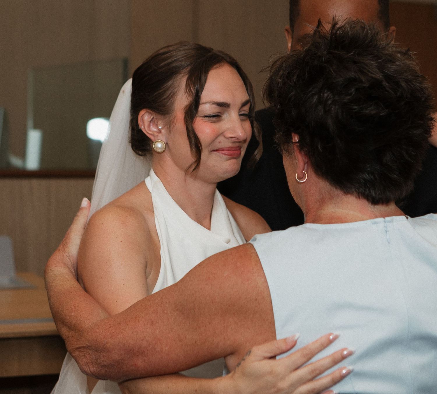 A bride in a white wedding dress shares an emotional embrace at an indoor venue.