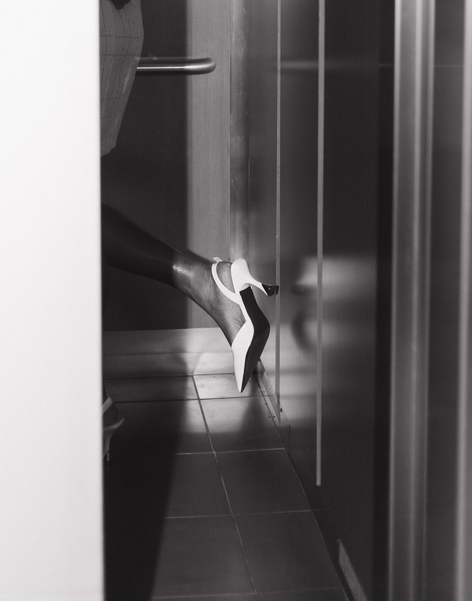 A black and white photograph shows a high heel shoe dramatically lit in a dark hallway.