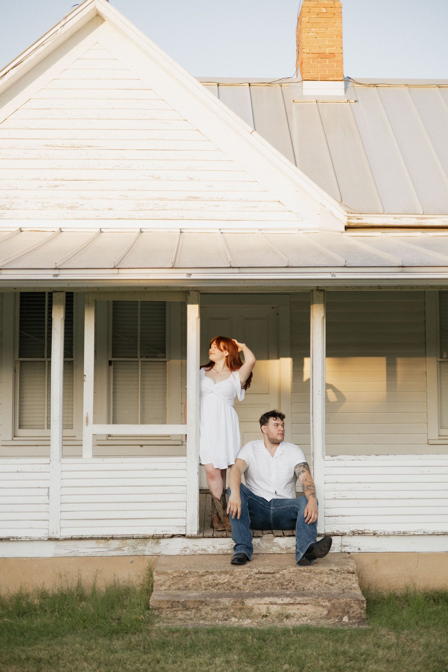 Couple in white outfits on porch of vintage white farmhouse, woman standing, man sitting on steps.