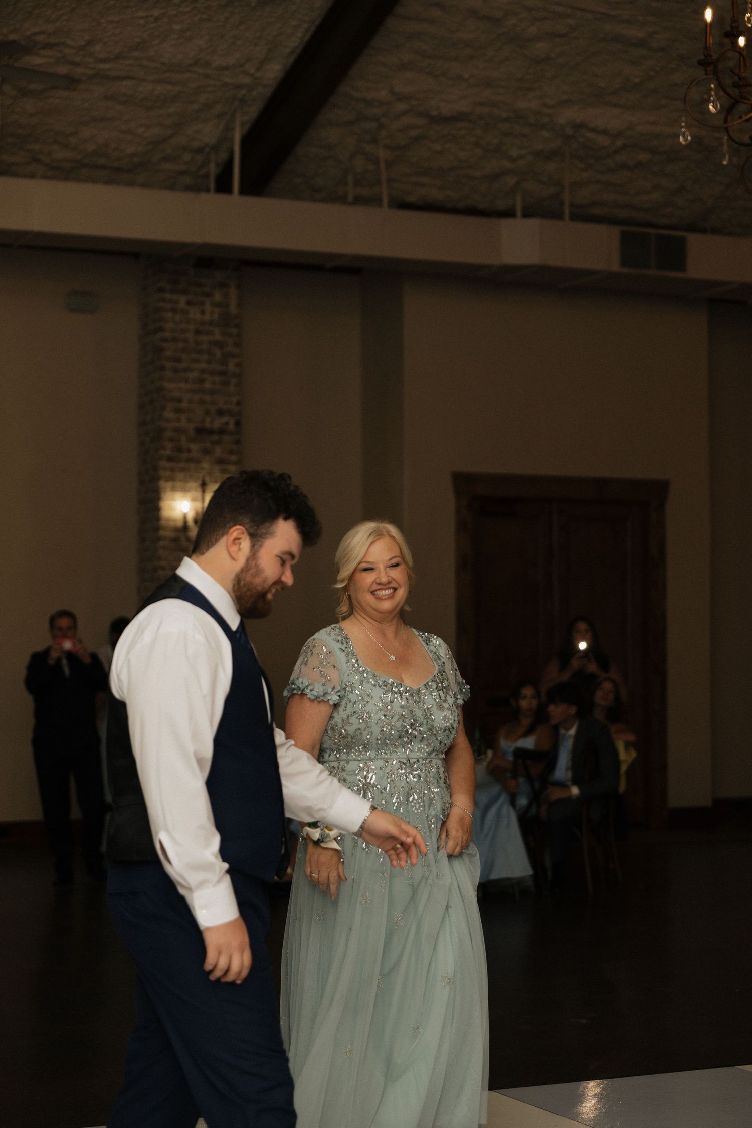 Groom in navy vest shares mother-son dance with smiling woman in silver sequin gown at reception.