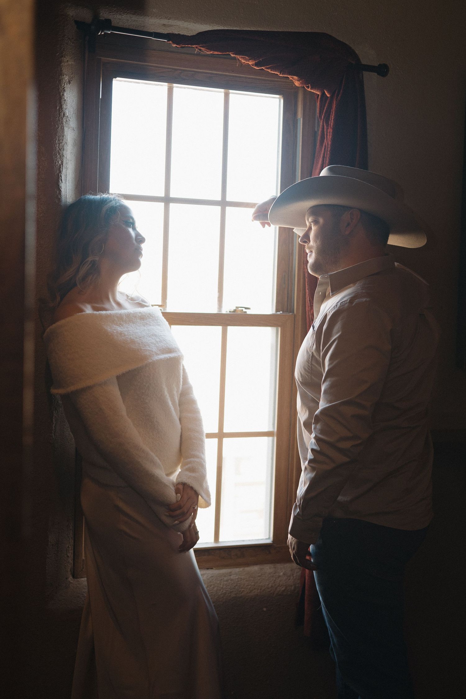 Bride and groom in cowboy hat share intimate moment by rustic window in warm backlit wedding portrait.