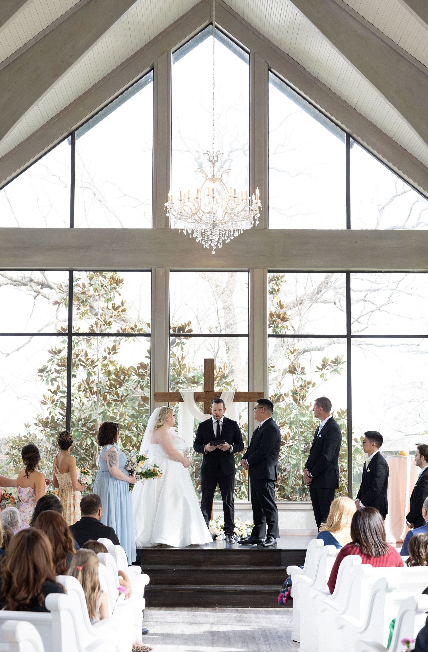 Elegant wedding ceremony in a chapel with vaulted ceilings, crystal chandelier, and large windows framing the couple at the altar.