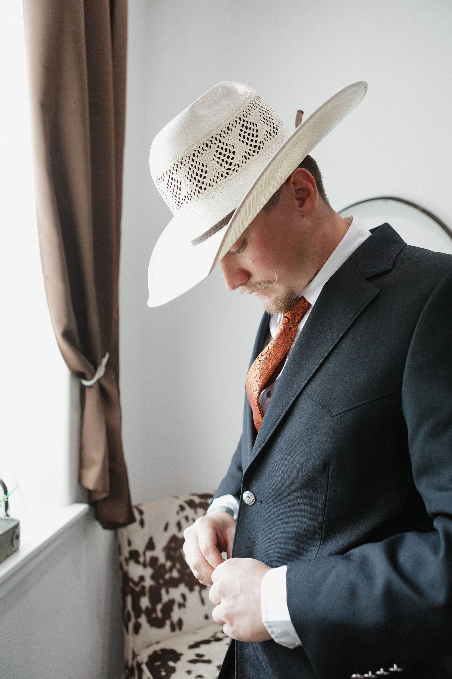 Person in cowboy hat and suit adjusts buttons while standing by window.