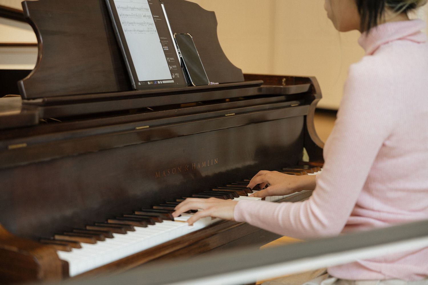 Young woman in pink sweater playing a grand piano with sheet music, seen from the side in a music room.