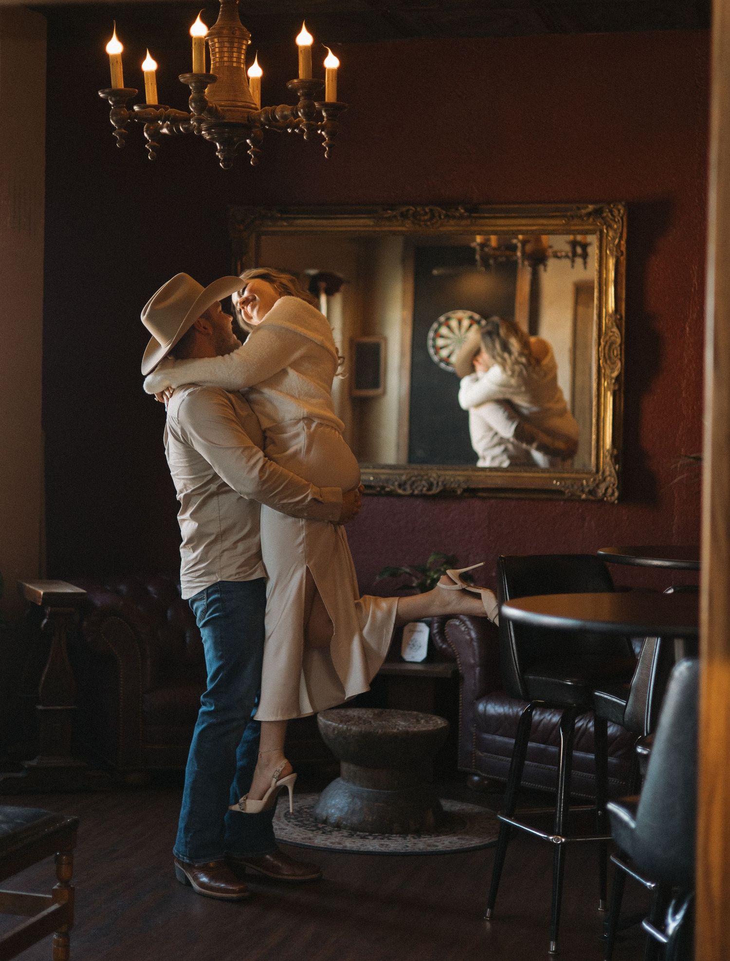A romantic embrace in a dimly lit room with an ornate chandelier and mirror on the wall.