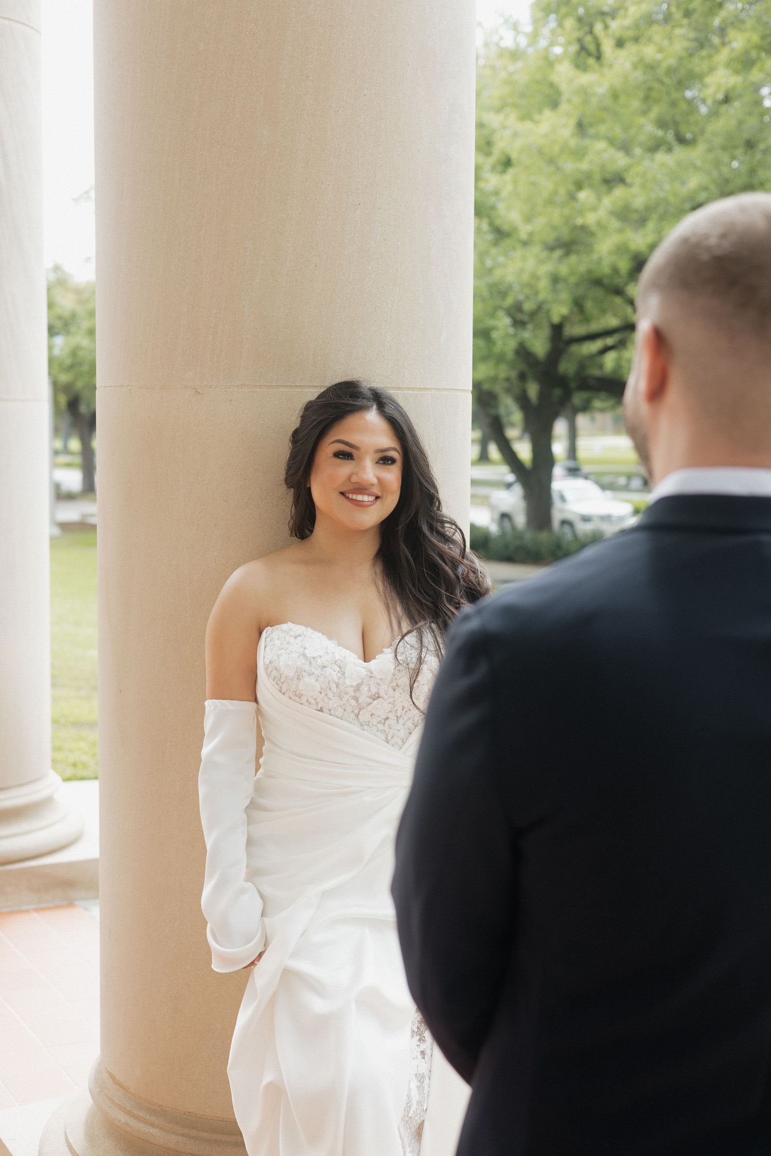 Bride in strapless lace wedding gown and white gloves leans against column, smiling at groom in black suit.