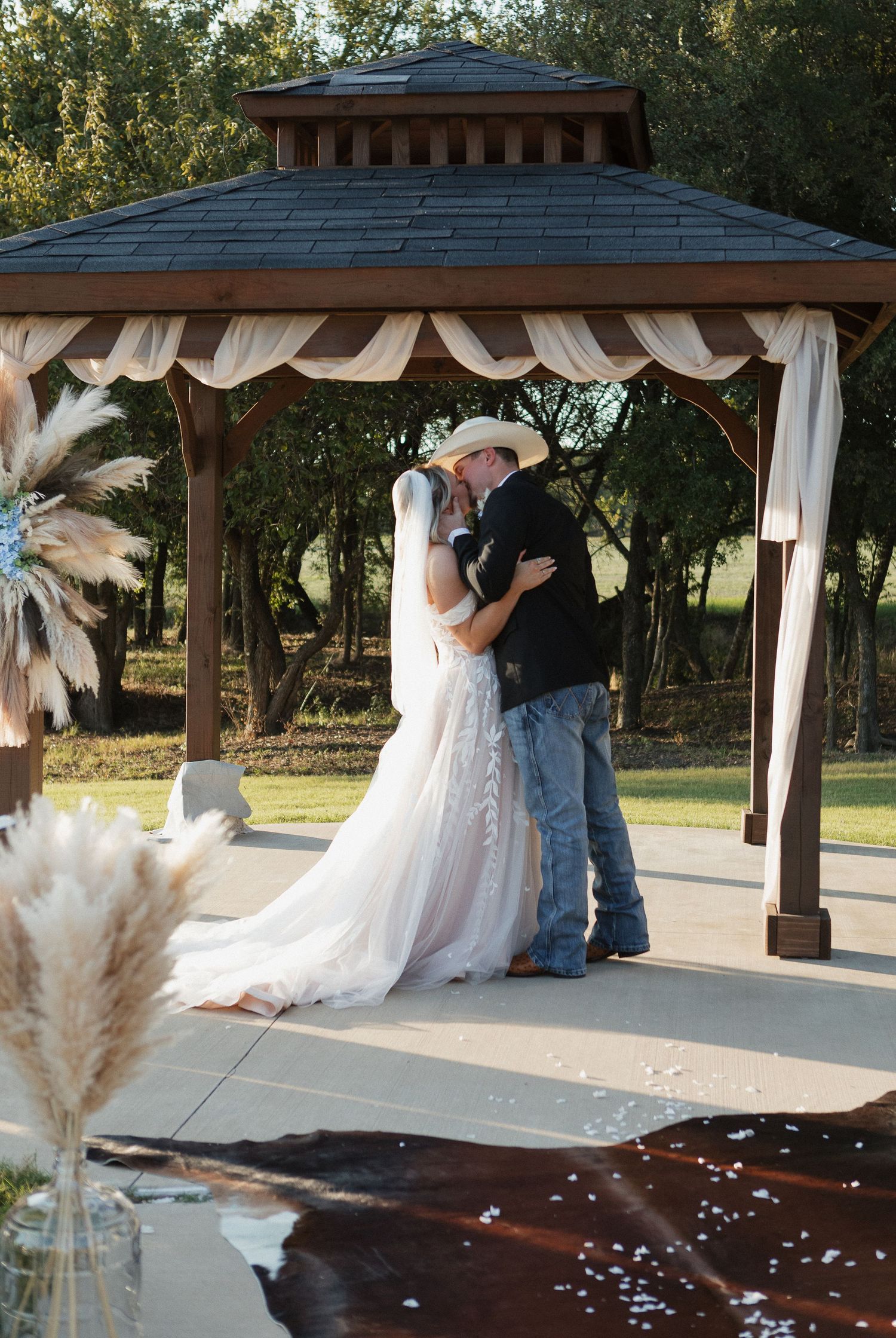 Bride and groom share first kiss under decorated gazebo at outdoor country wedding ceremony.