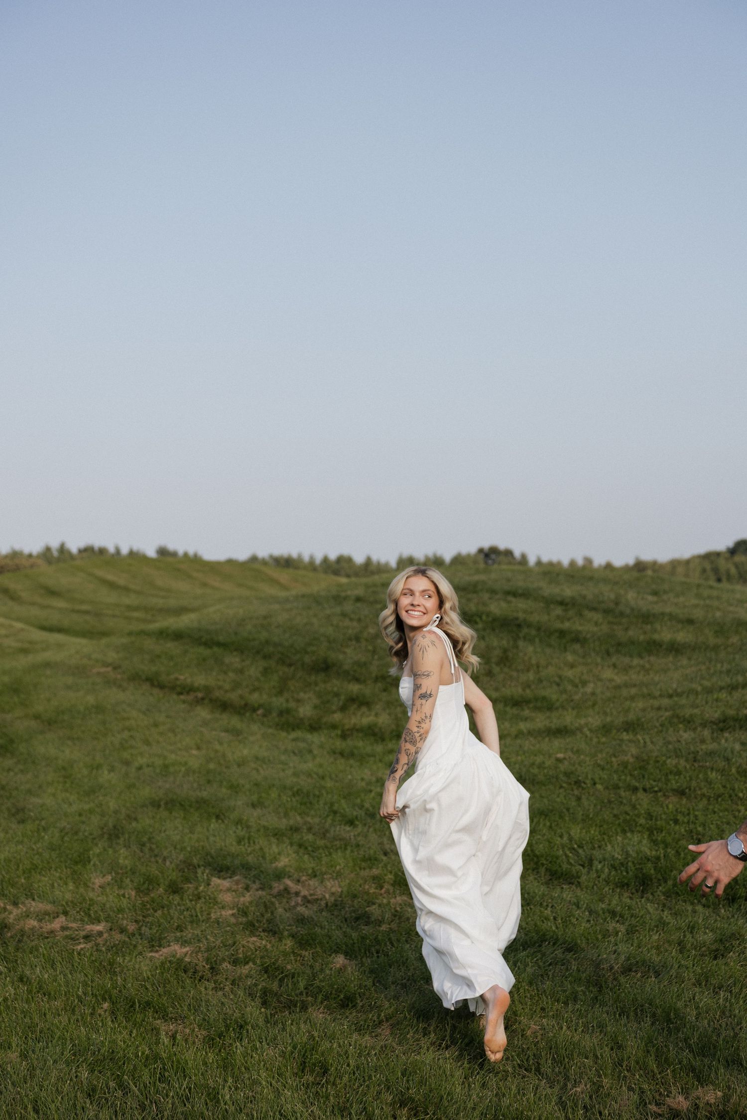 Smiling bride in white dress running through lush green field under clear blue sky.