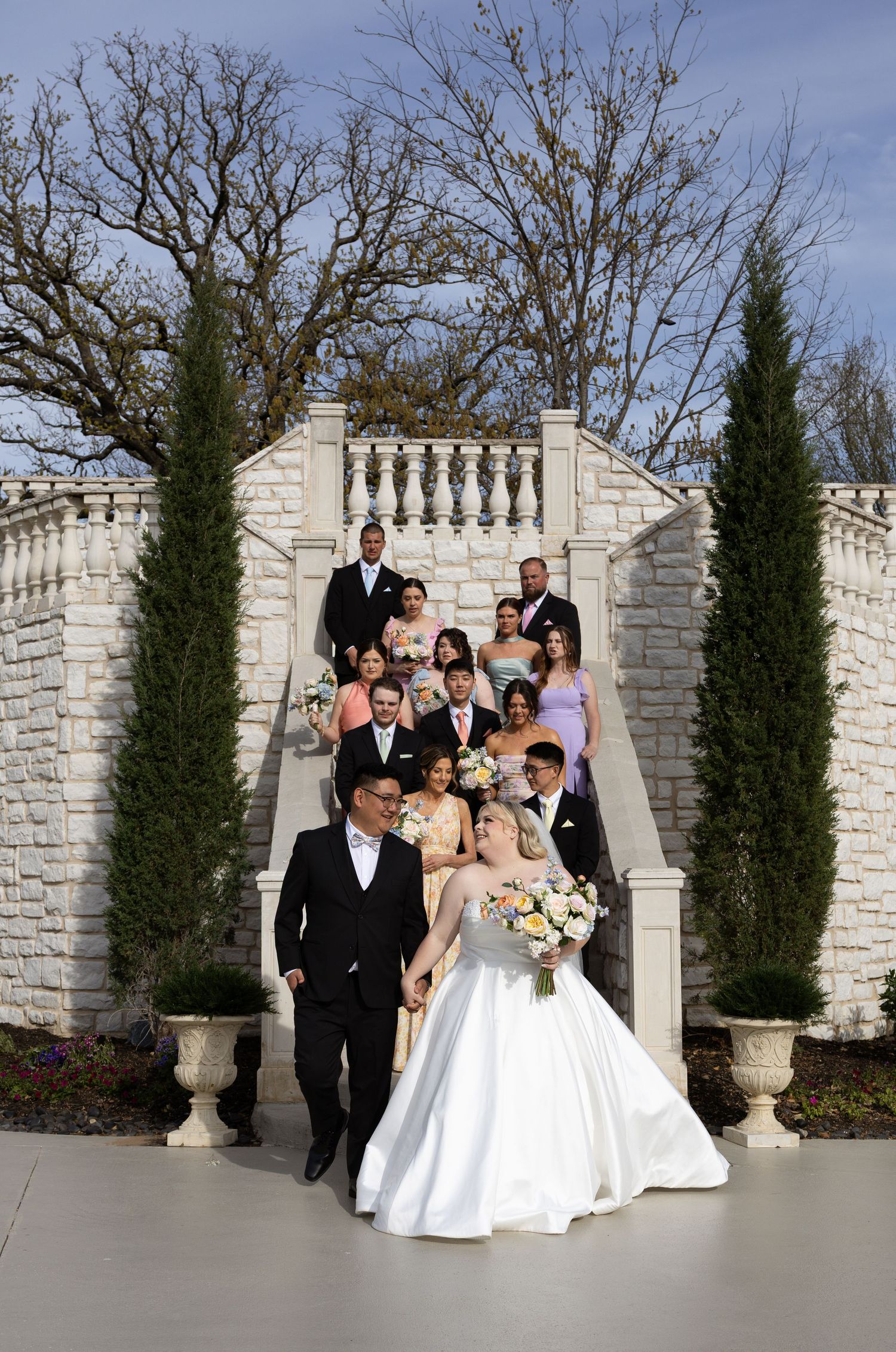 Wedding party posing on stone staircase with bride and groom in foreground at outdoor venue.