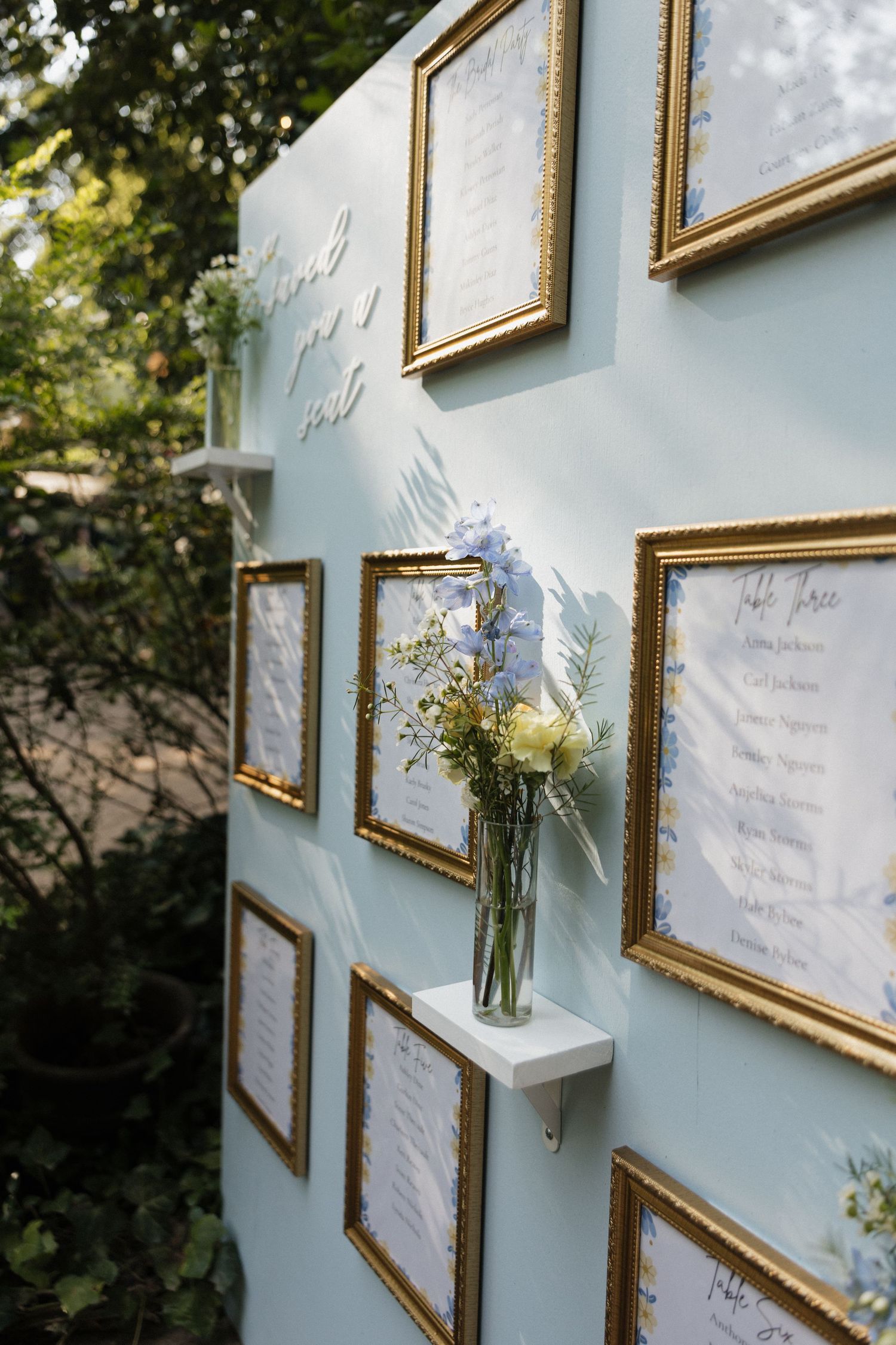 Gold-framed wedding seating chart display on a light blue wall with fresh flowers in glass vases.