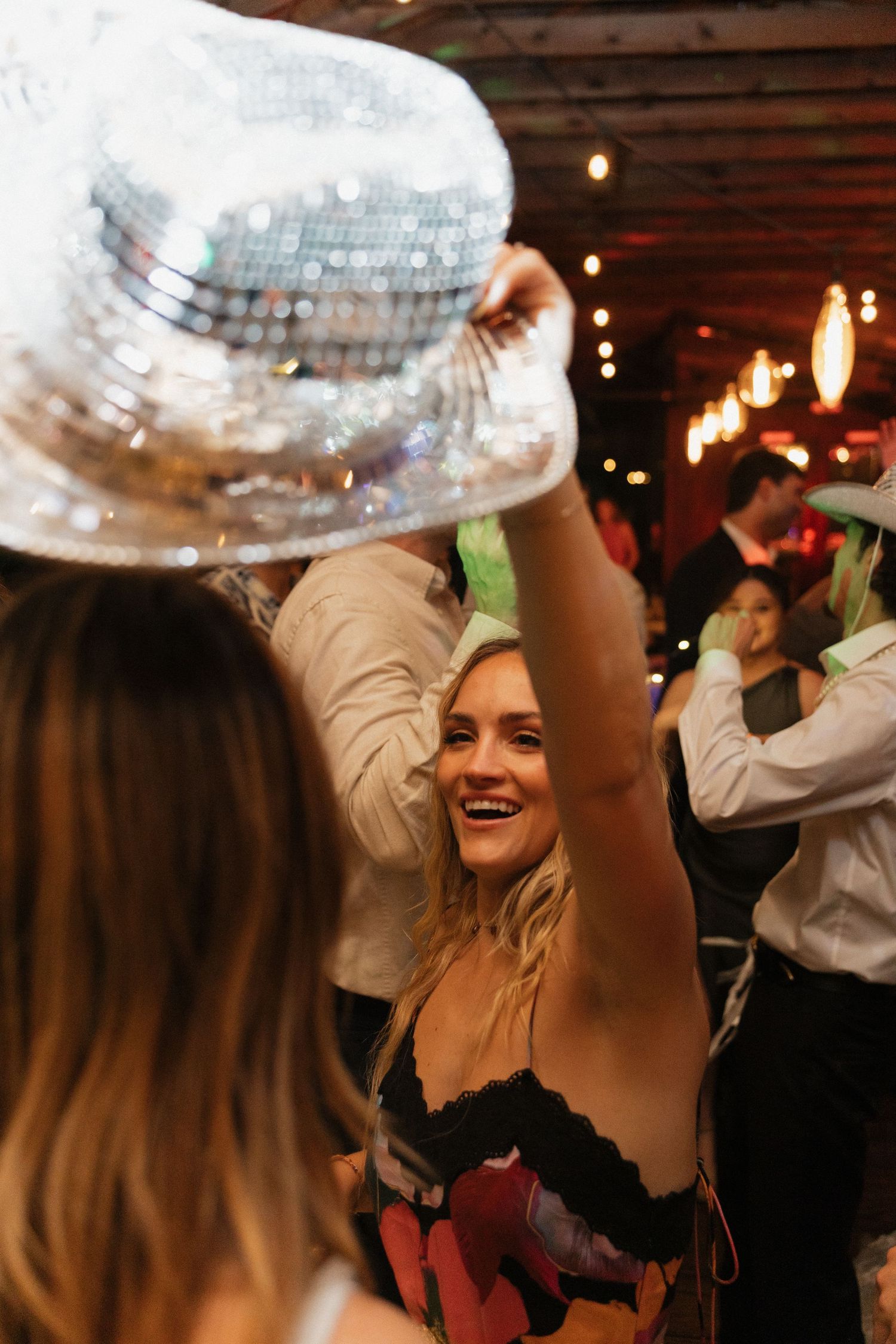 Woman smiling at a party holding a silver sequin disco ball hat under warm string lights. Caddo Mills Texas wedding photographer