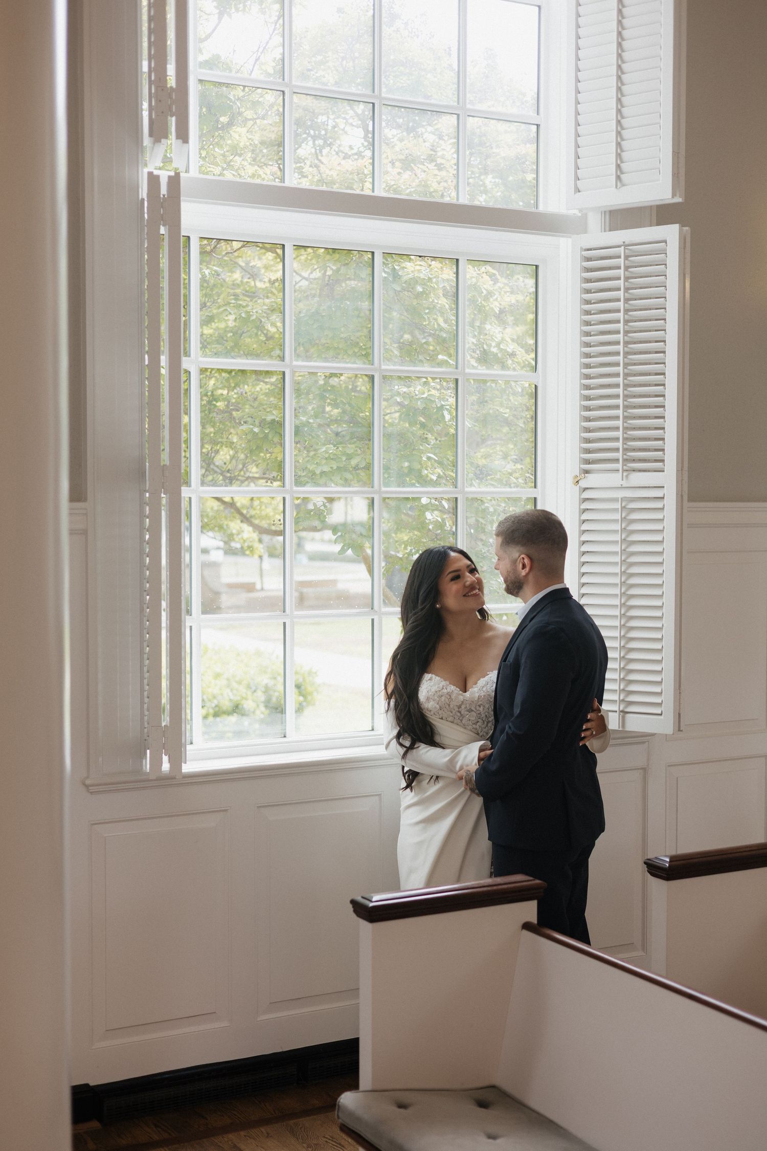 Bride and groom sharing a romantic moment by a large window on a staircase indoors.