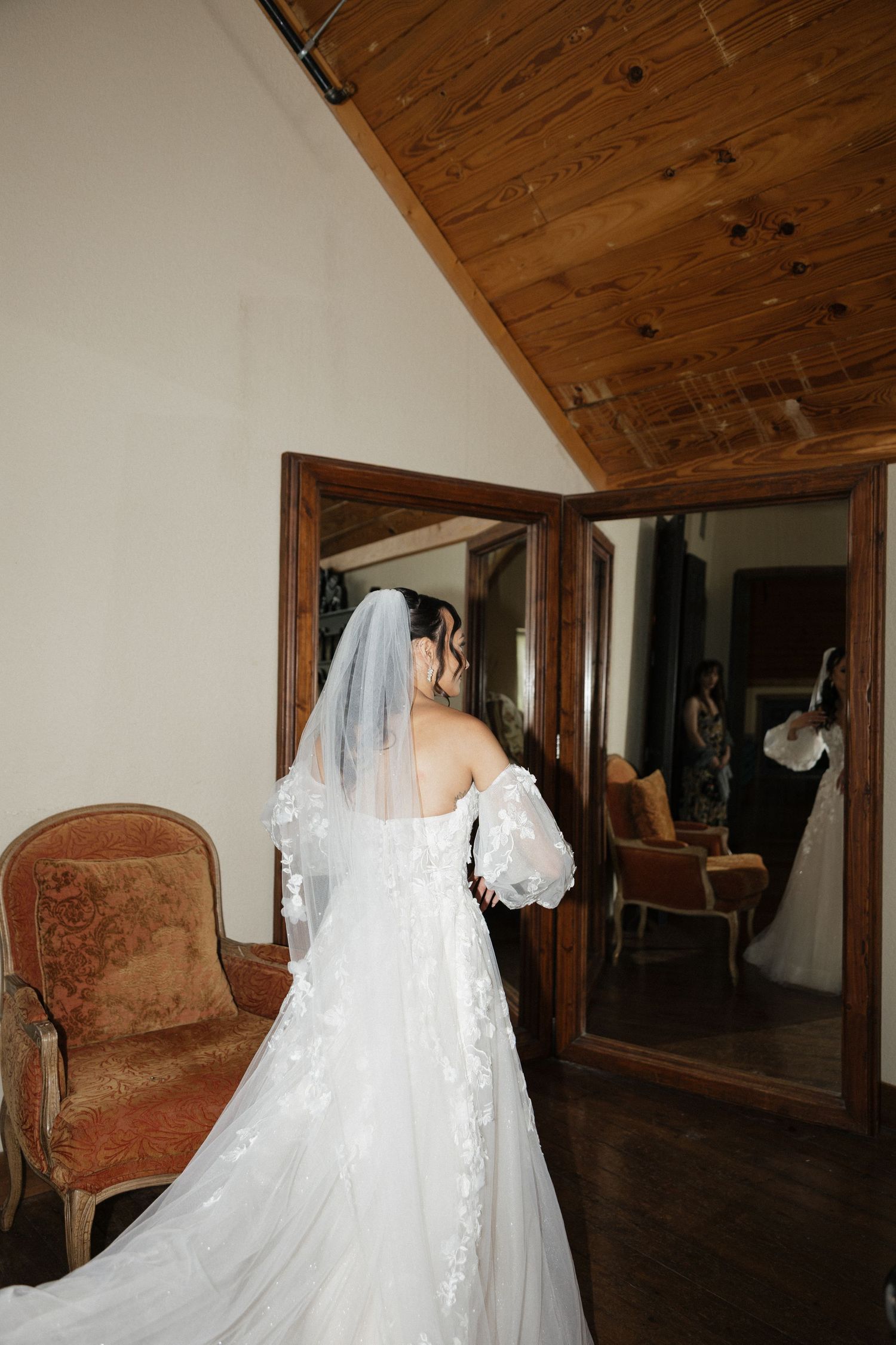 Bride in off-shoulder lace gown and veil views reflection in ornate mirror in rustic wood-ceiling room.