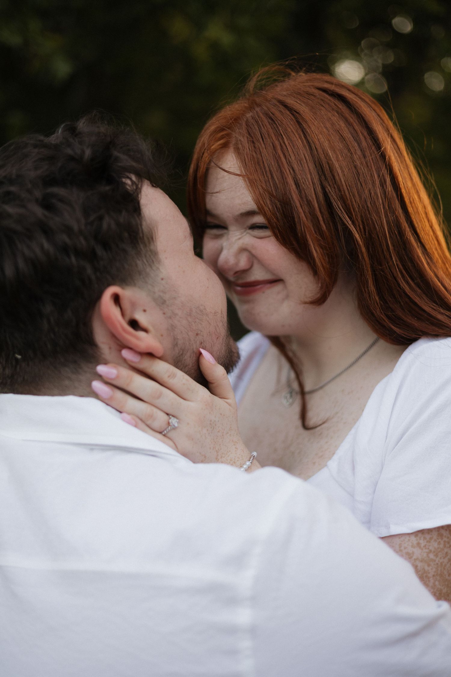 A smiling redhead woman with freckles leans in to kiss a dark-haired man outdoors, wearing a diamond ring.