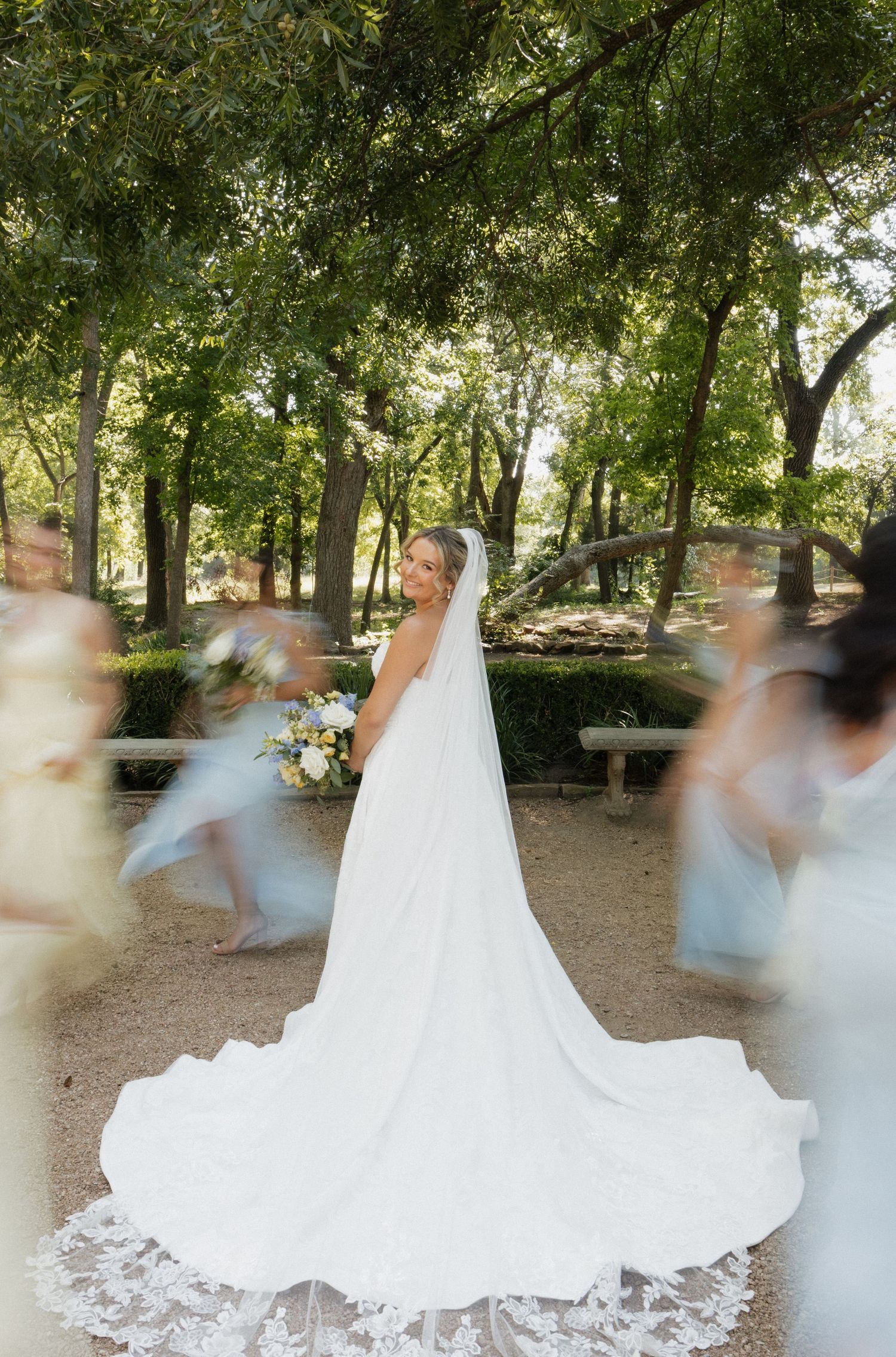 Bride in white gown and veil stands still as bridesmaids blur around her in an outdoor garden setting.