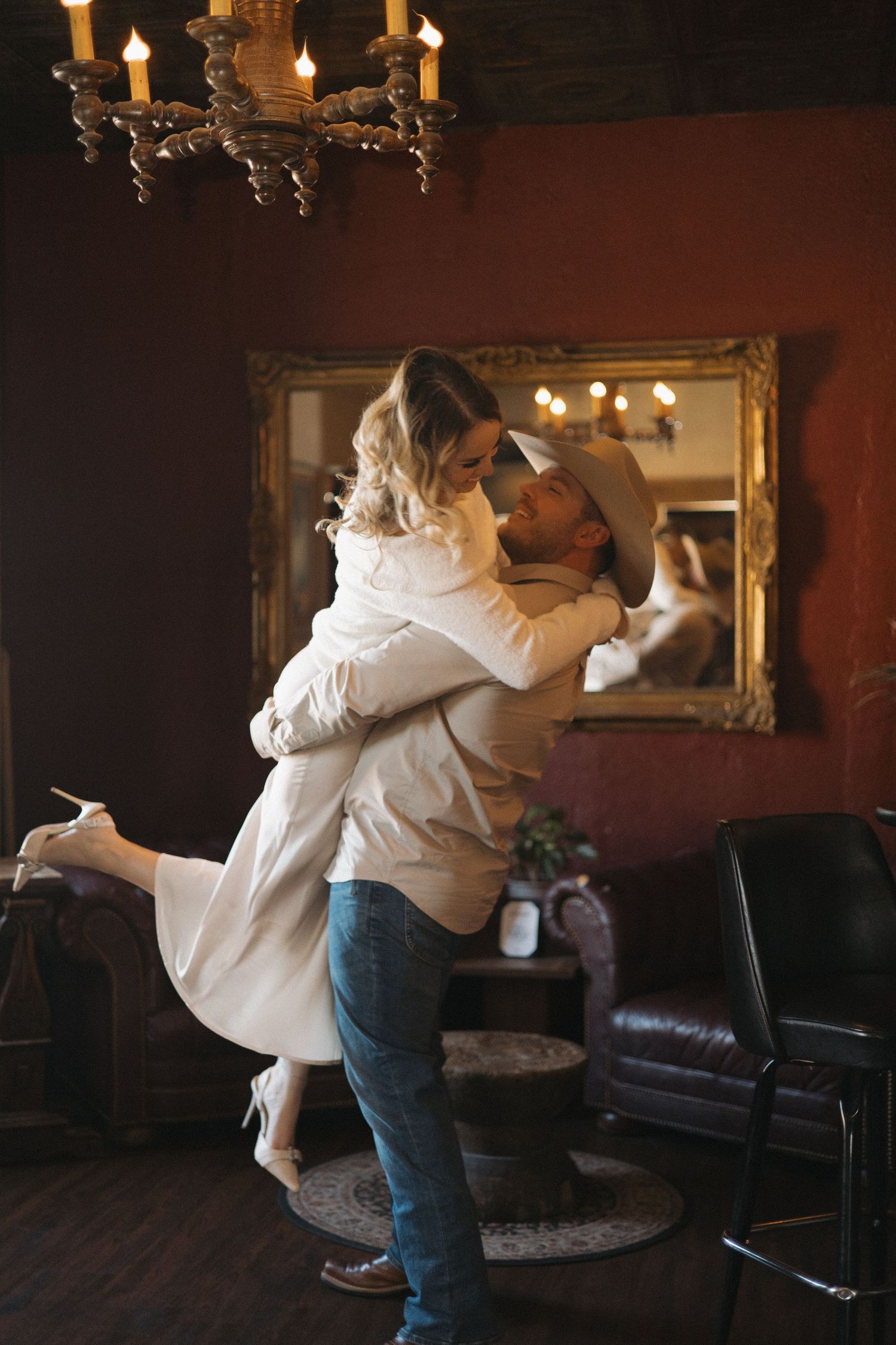 Couple sharing romantic embrace in rustic decorated room with chandelier and vintage mirror, man lifting woman in wedding attire.
