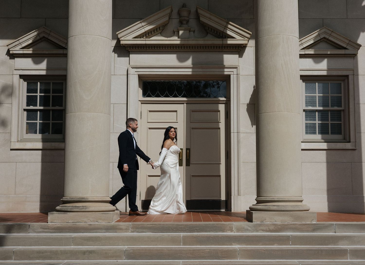 A bride and groom hold hands on steps of an elegant columned building during their wedding day photoshoot.