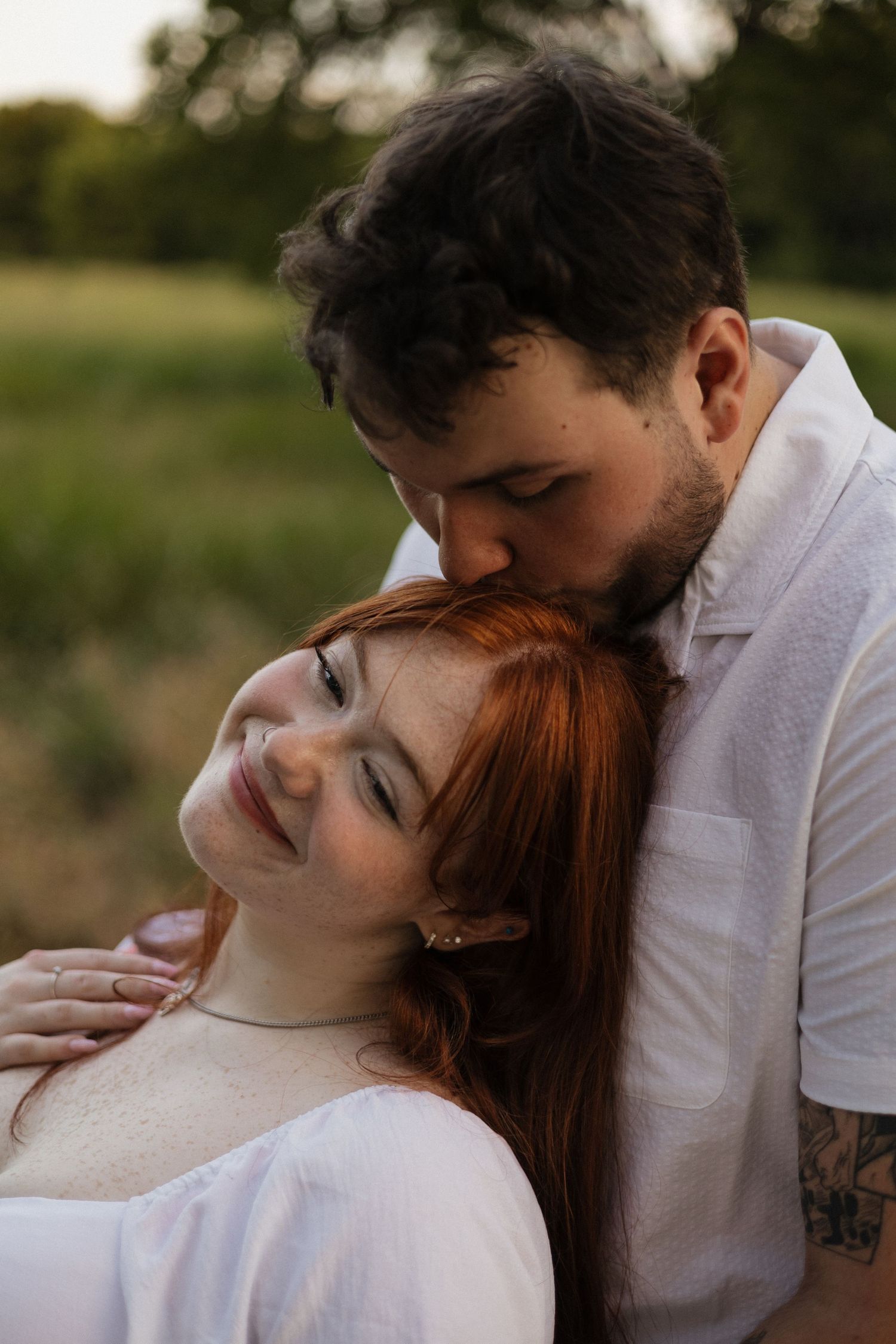 A man with dark curly hair kisses a smiling redhead woman outdoors in a green field setting.