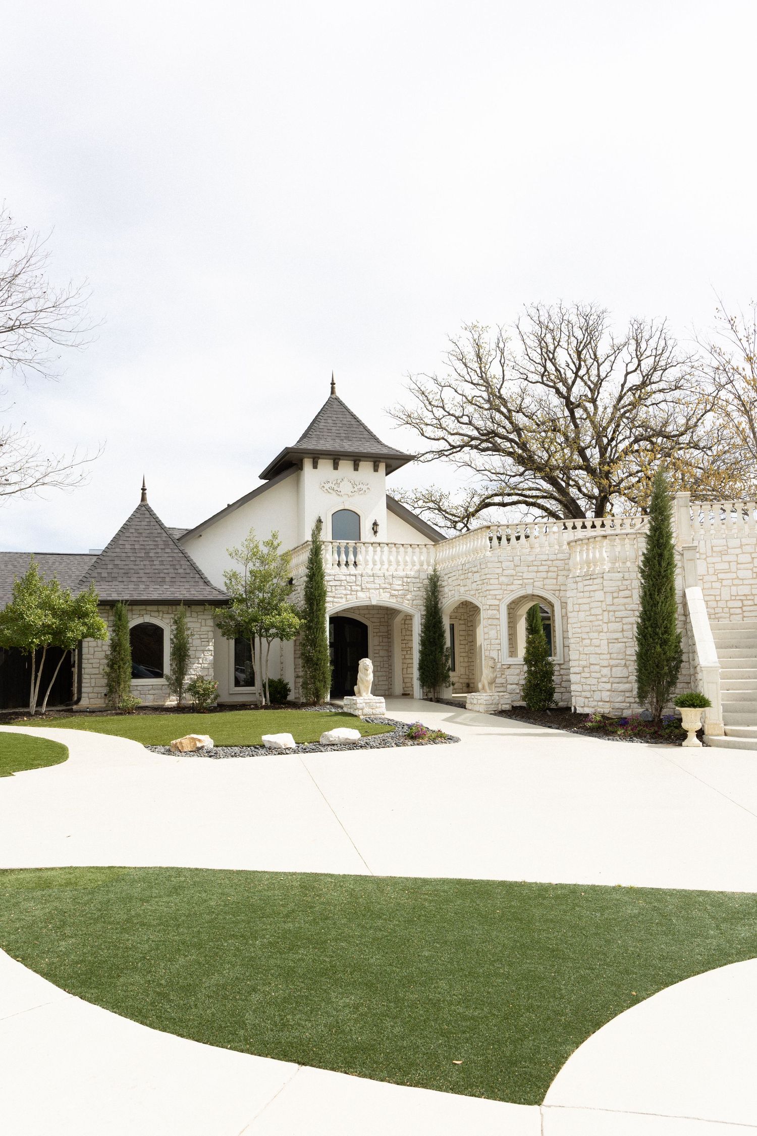 Elegant castle-style mansion with stone walls, turret, arched entry, cypress trees, and manicured lawn on a wide concrete driveway.