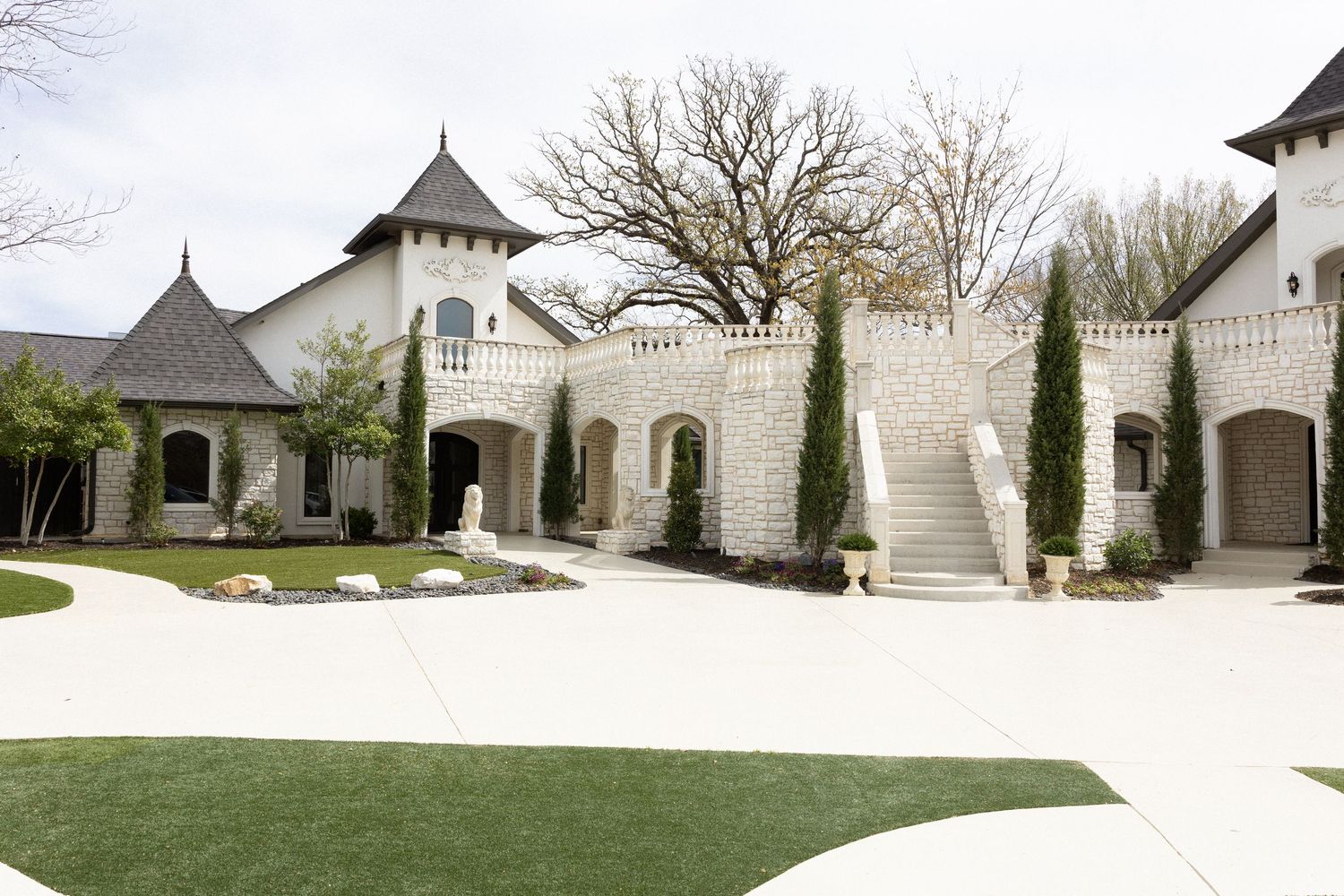 Grand castle-style mansion with white stone walls, turrets, arched entries, cypress trees, and a sweeping driveway.
