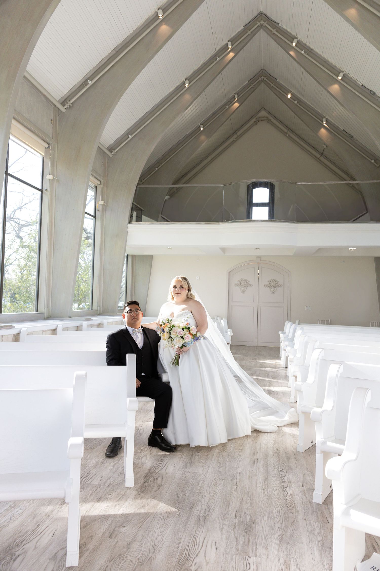 Bride in white gown and groom in black suit pose in a bright white chapel with vaulted ceilings and wooden pews.