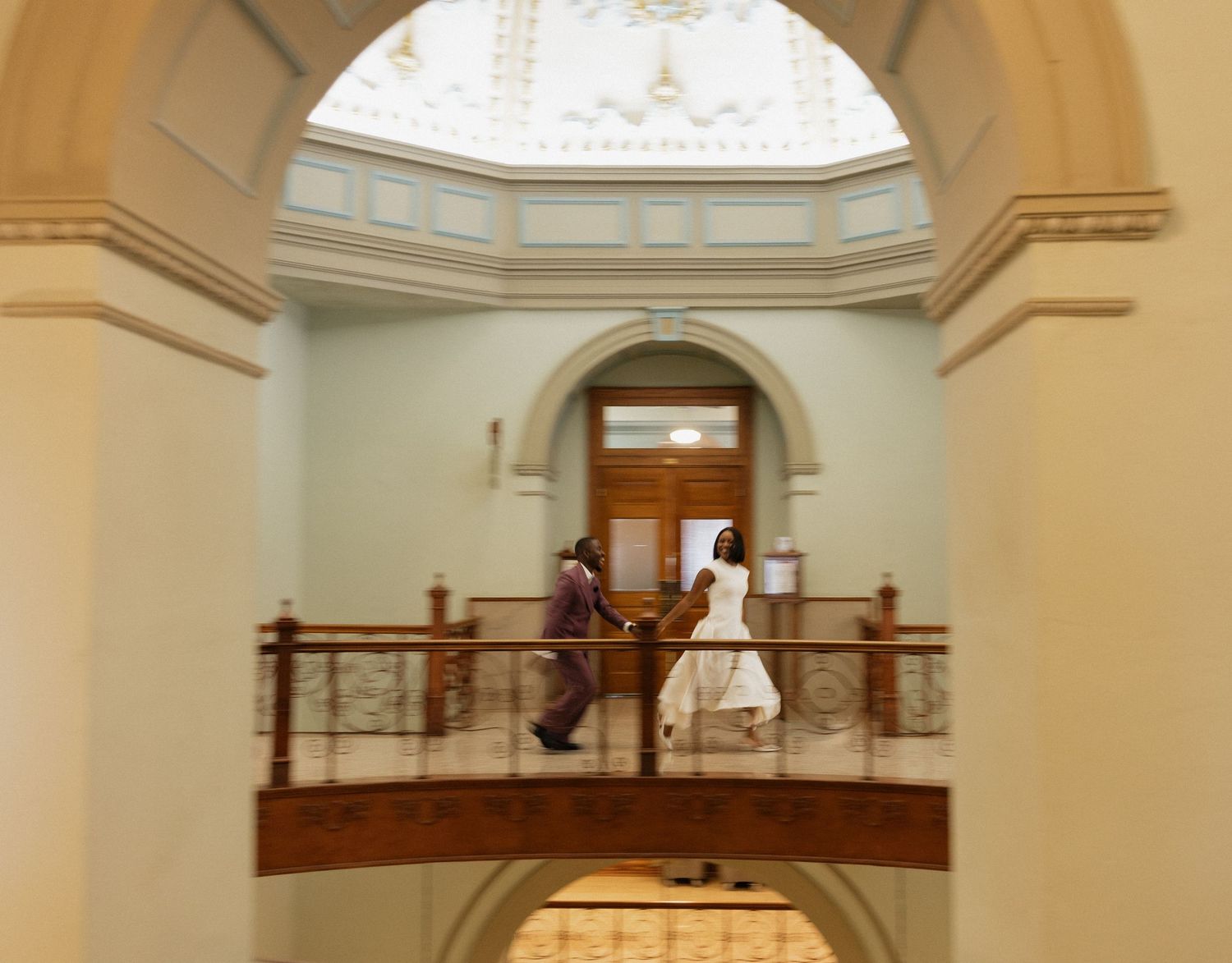 Bride and groom walking across an elegant indoor balcony with arched ceilings in a historic building.