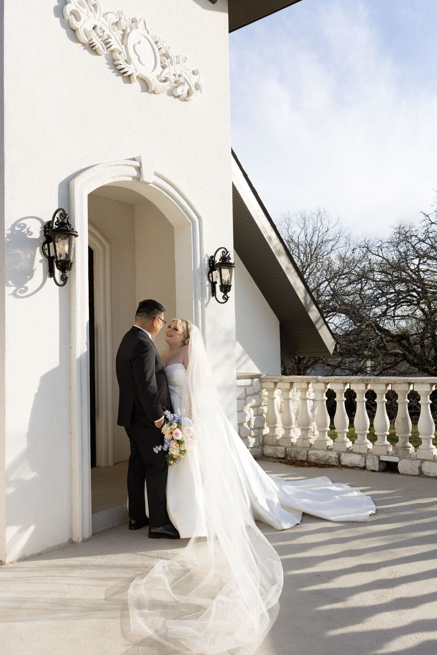 Bride and groom share a romantic moment in an elegant arched doorway, wedding veil flowing on a sunlit balcony.