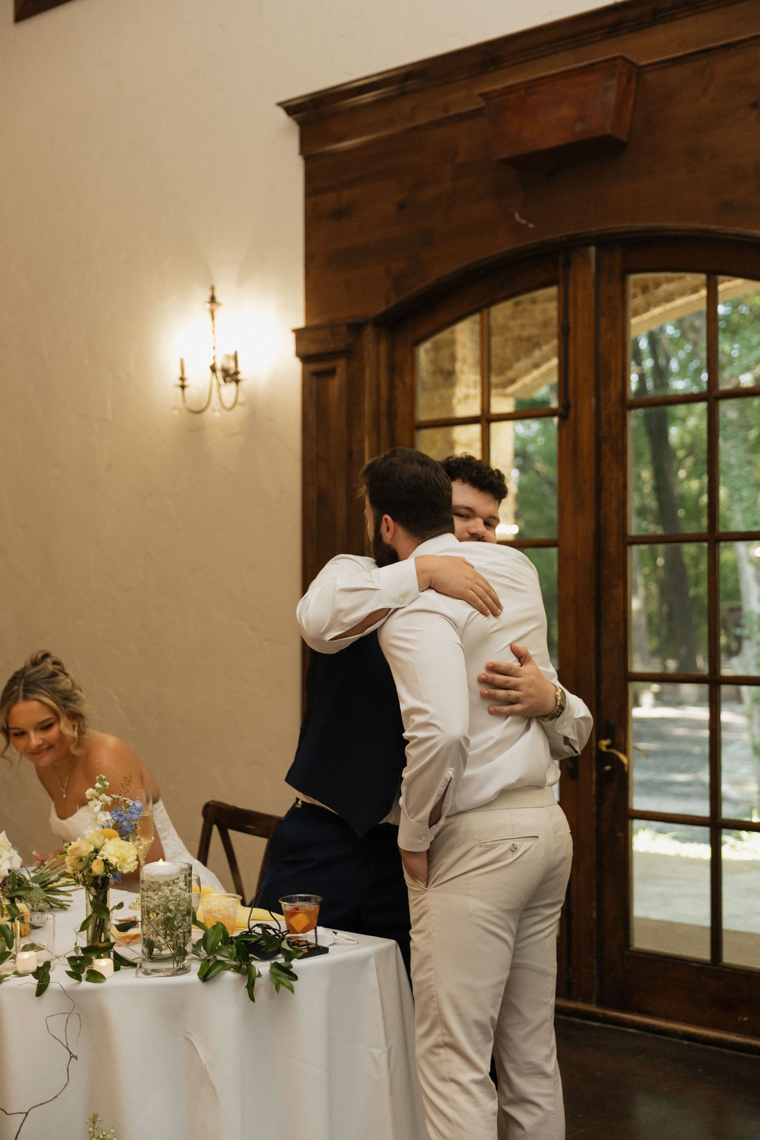 Two grooms share an emotional hug at their wedding reception near a decorated table with floral arrangements.