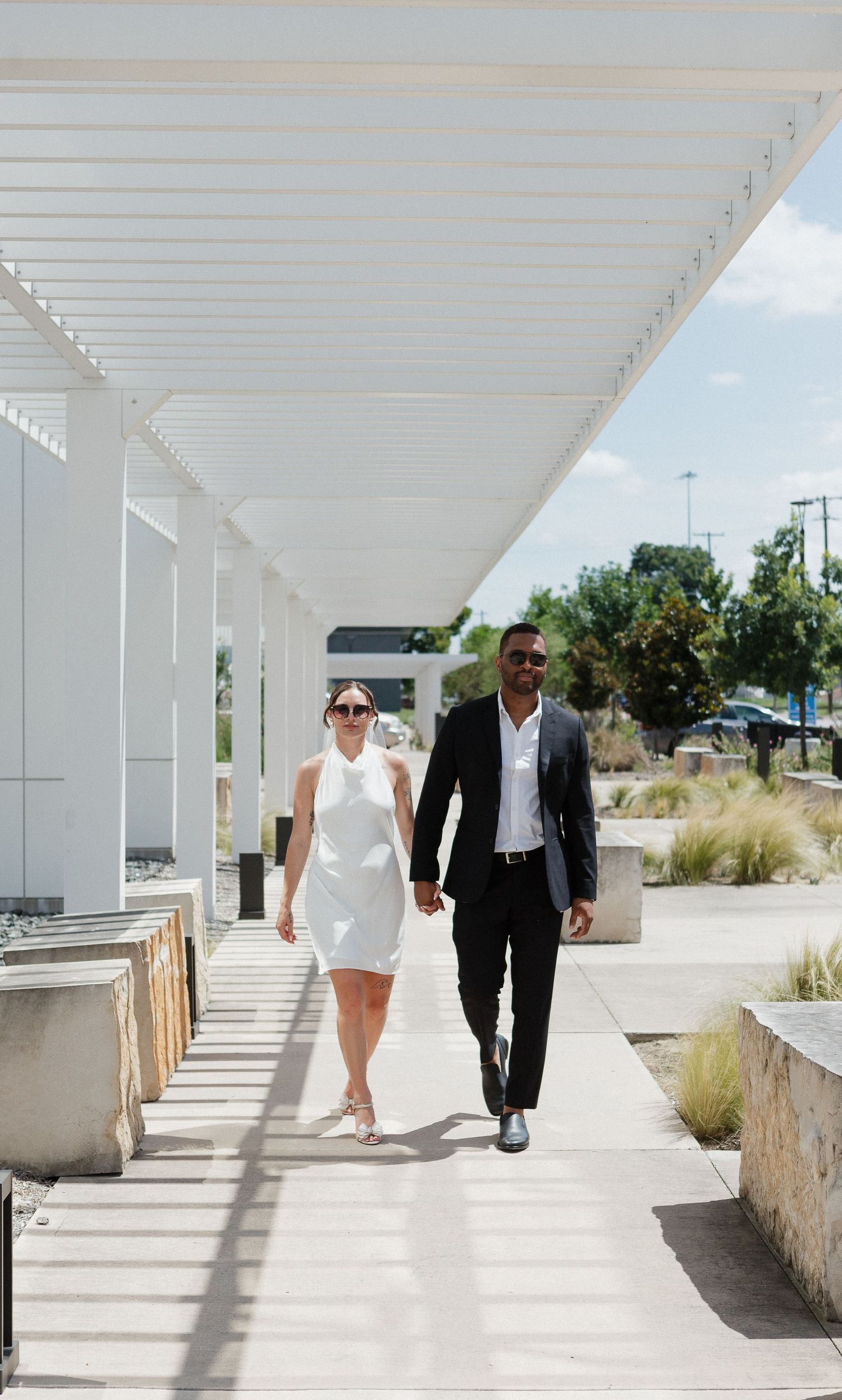 Two people walk together along a bright white covered walkway with stone walls and coastal vegetation visible outside.