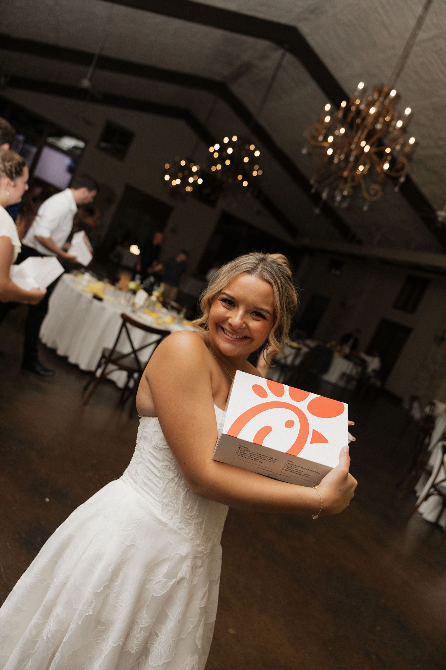 Smiling bride in white wedding gown hugging a Chick-fil-A box at her elegant reception venue.