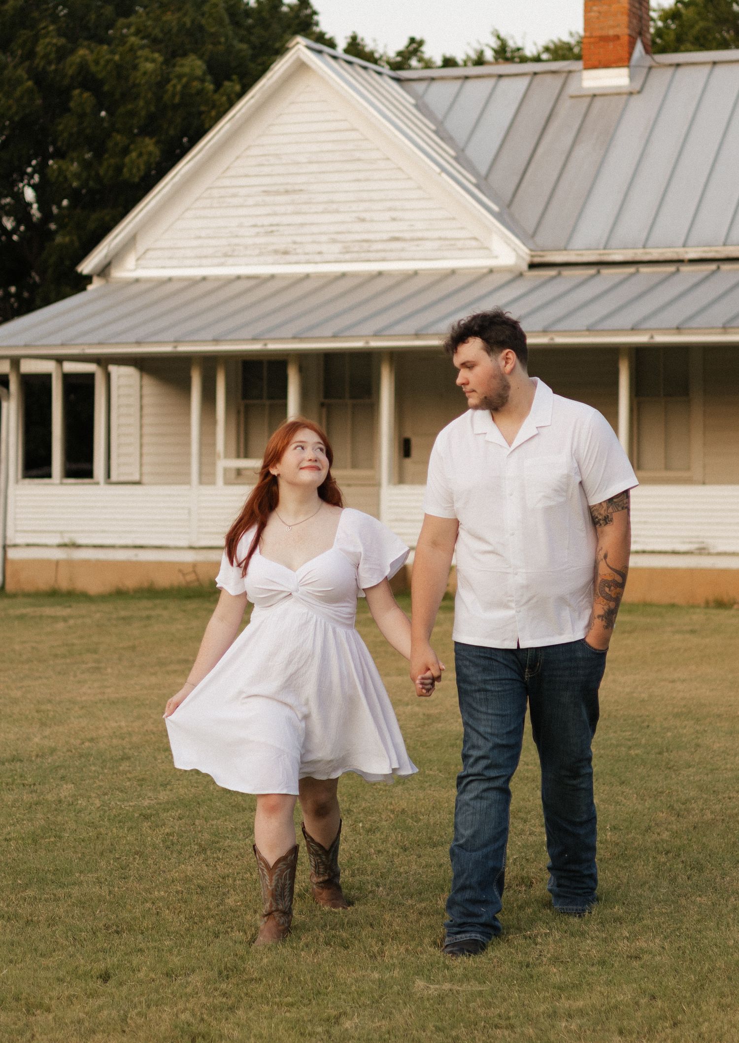 A couple in white outfits holds hands walking on grass before a charming farmhouse at golden hour.