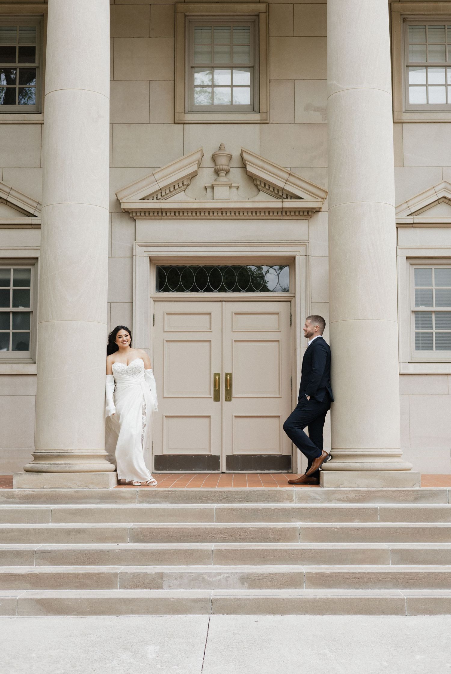 Bride and groom pose on steps of elegant columned building in wedding attire.
