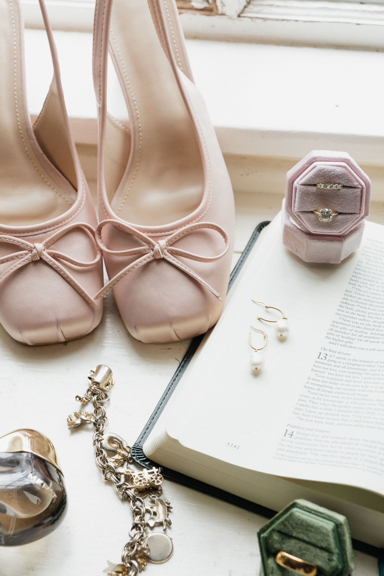 Blush pink bow heels beside an open Bible, pearl earrings, charm bracelet, and a diamond ring in a velvet box.