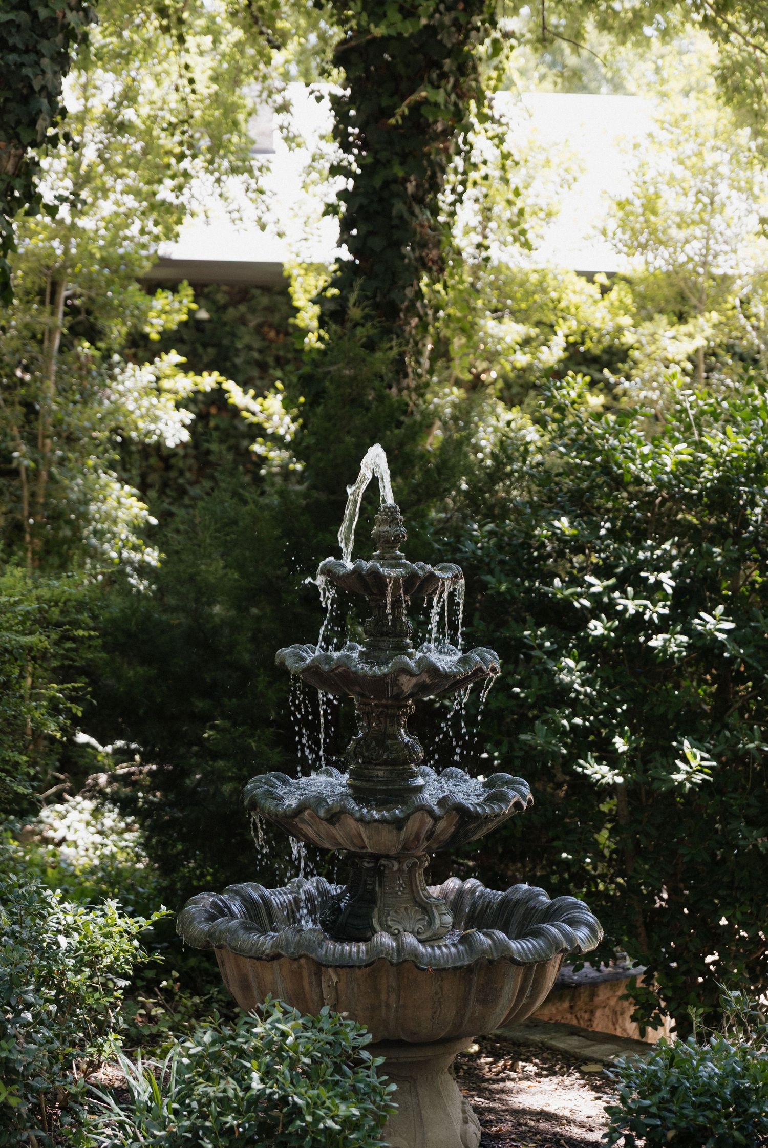 A elegant three-tiered stone garden fountain with cascading water, surrounded by lush green trees and ivy-covered walls.