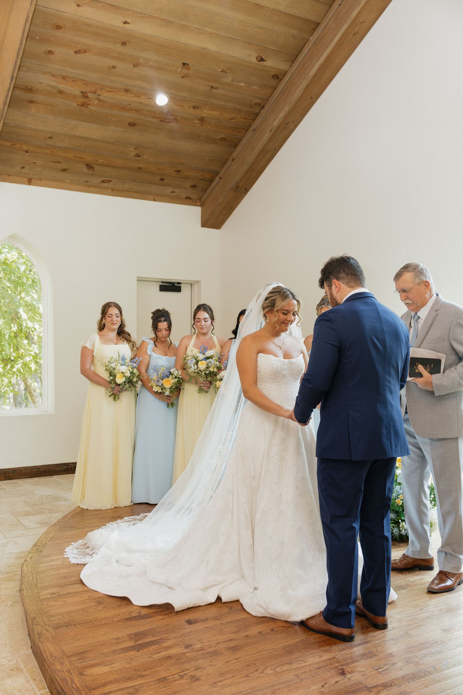 Bride and groom hold hands at altar with bridesmaids in yellow and blue dresses during ceremony.