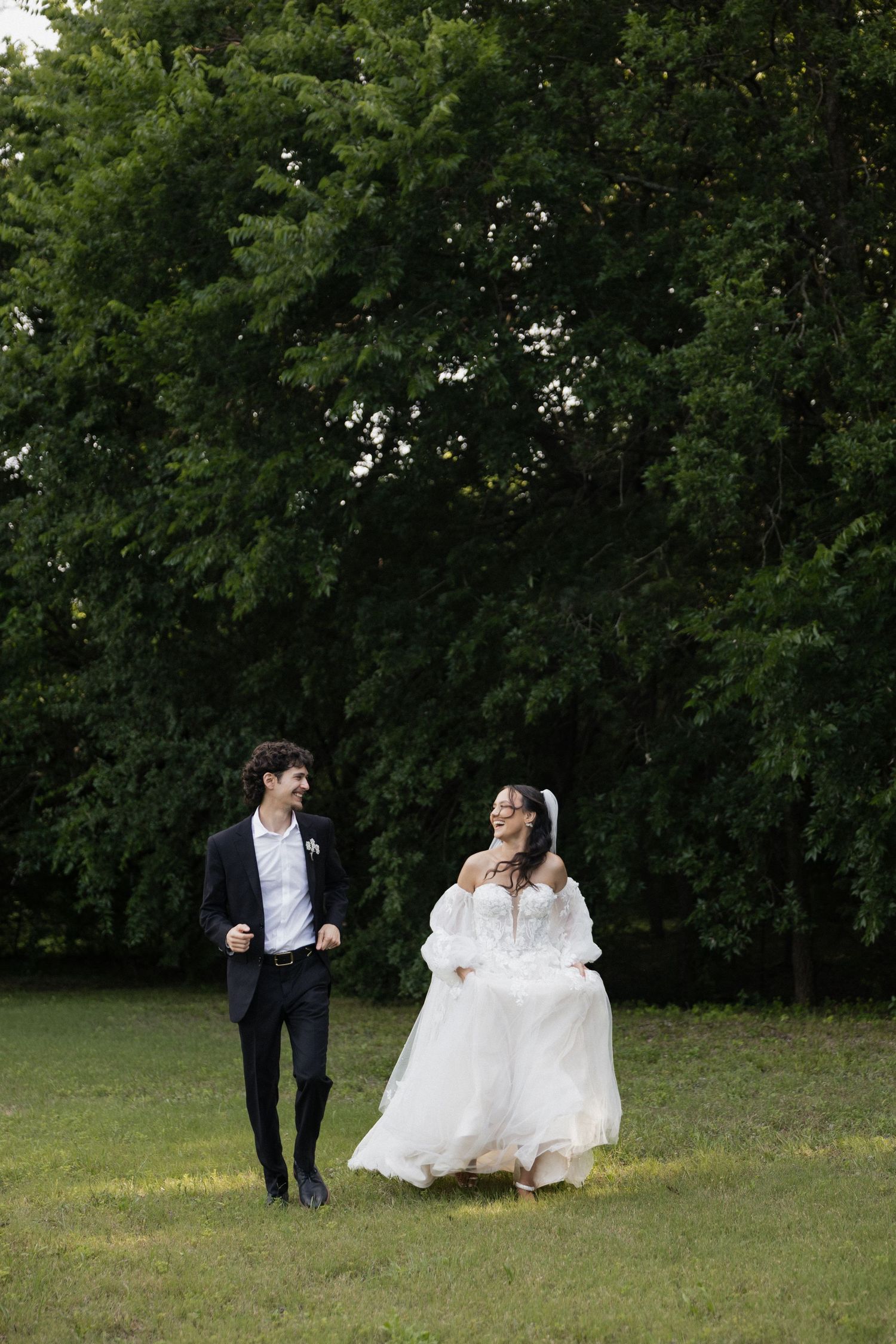Happy bride in white gown and groom in black suit walk together on green grass with trees behind them.