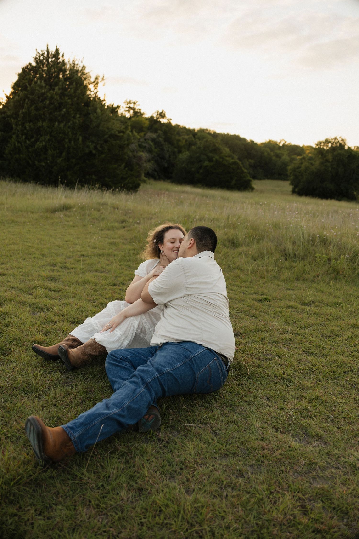 Couple sitting in a grassy field at sunset, embracing and kissing, wearing jeans and white tops with cowboy boots.