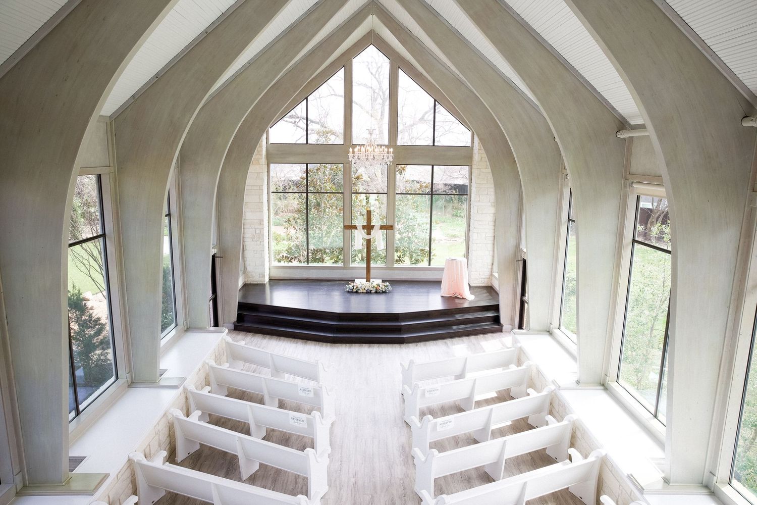 Elegant white chapel interior with arched windows, wooden altar, and rows of white pews bathed in natural light.