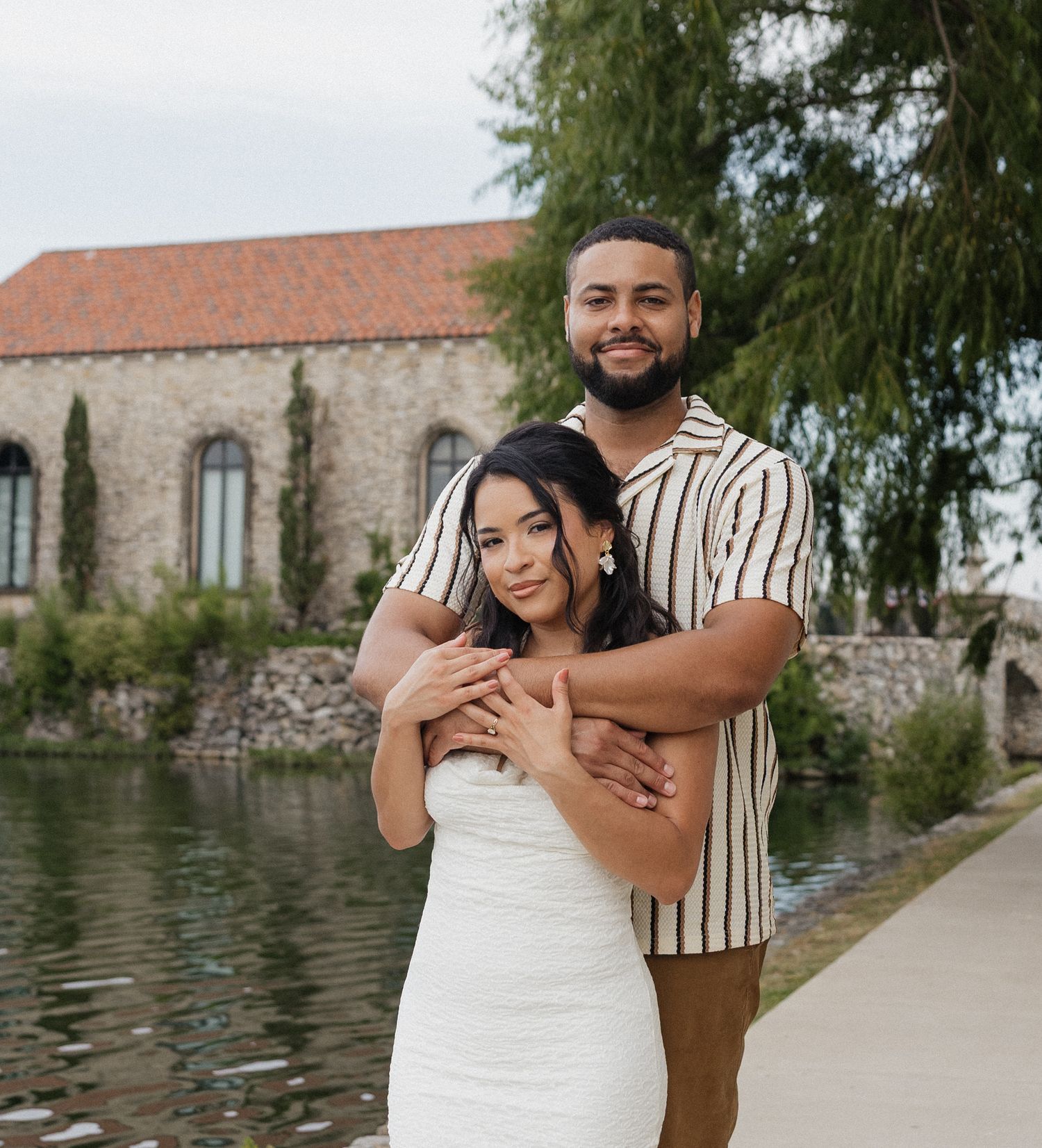 Couple posing romantically by a scenic waterway with a stone building and lush greenery in the background.
