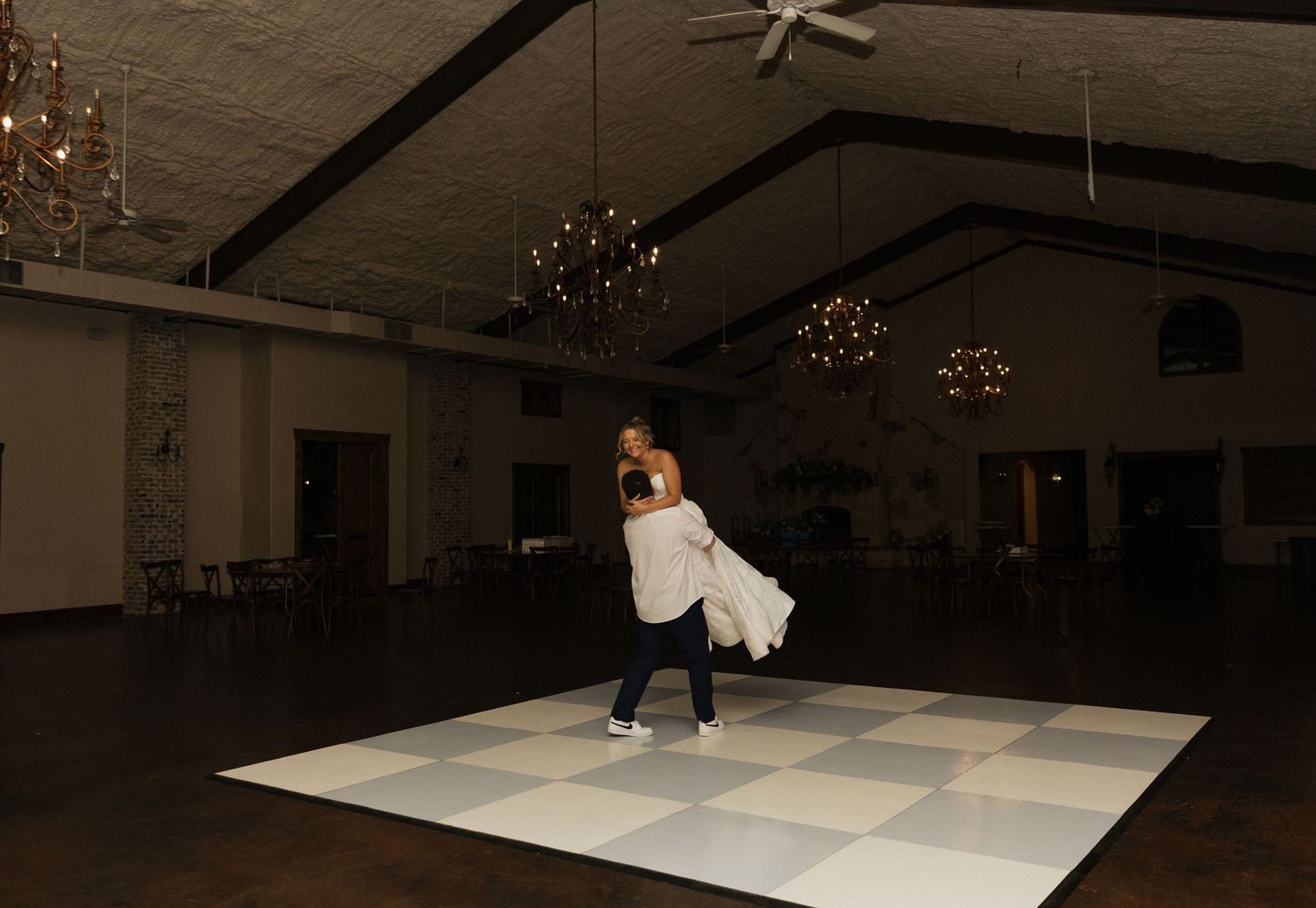 Couple sharing first dance on checkered dance floor in elegant ballroom with chandeliers at wedding reception.