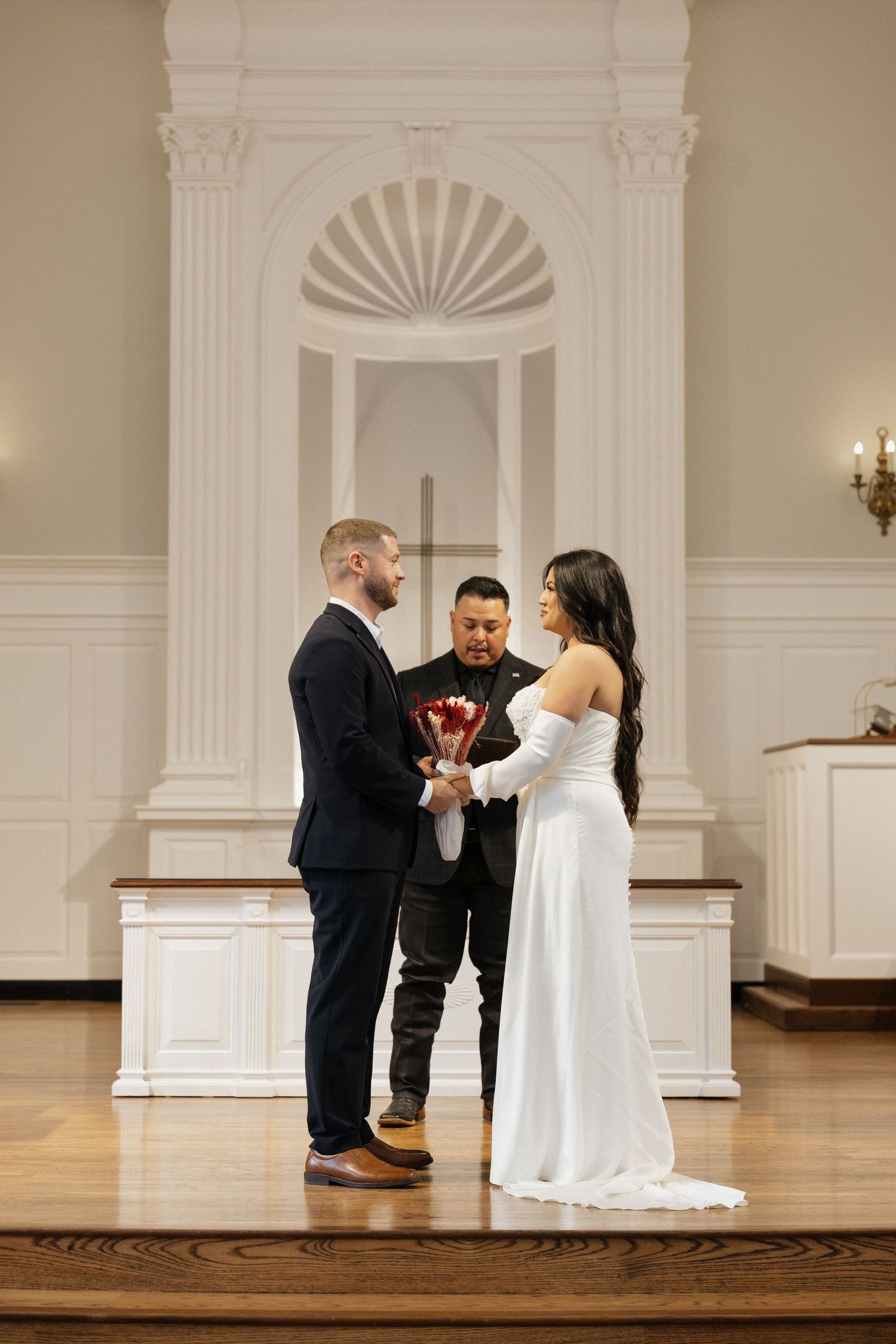 Couple exchanging vows at church altar, bride in white off-shoulder gown holding red roses, groom in navy suit.