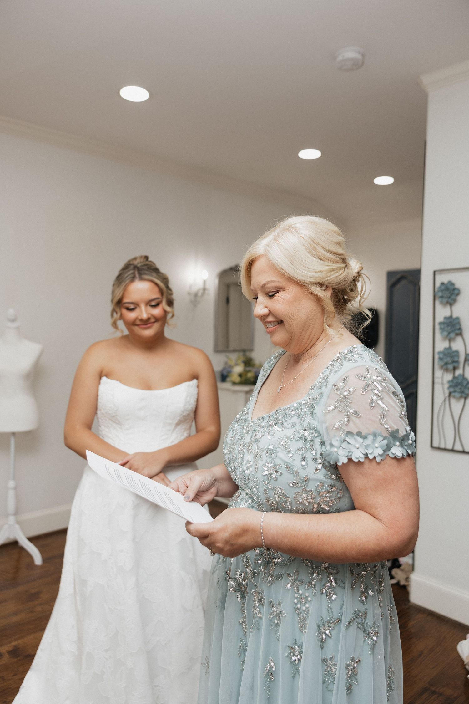 Bride in strapless white gown smiles as older woman in blue beaded dress reads a letter in elegant bridal suite.