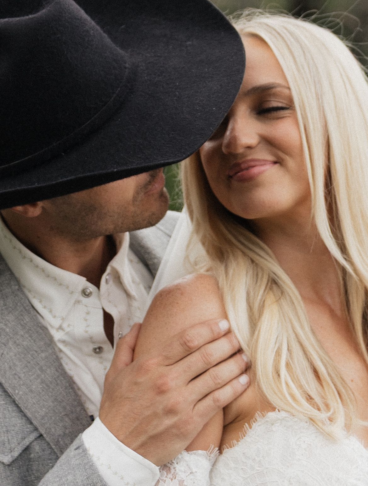 A couple shares a romantic moment, man in black cowboy hat kissing blond woman in white lace dress outdoors.