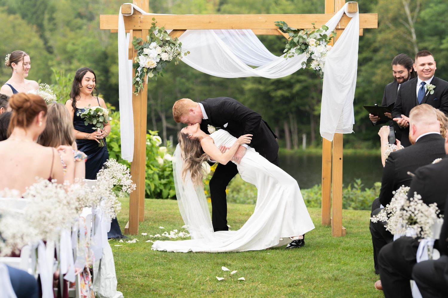 Bride and groom share a romantic dip kiss at outdoor wedding ceremony under wooden arch with white draping.