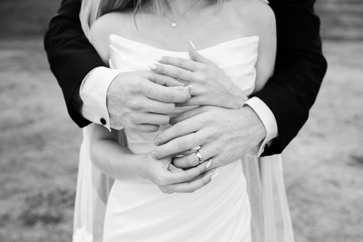 Black and white photo of bride and groom embracing, showing wedding rings on clasped hands.