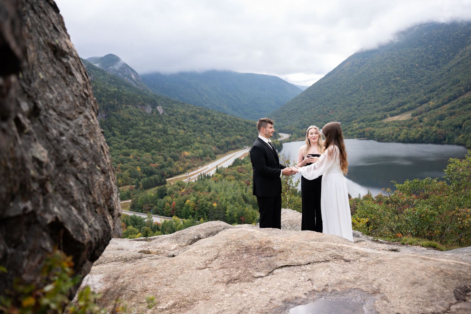 Couple exchanges vows with officiant on rocky mountain overlook with scenic lake and valley views.