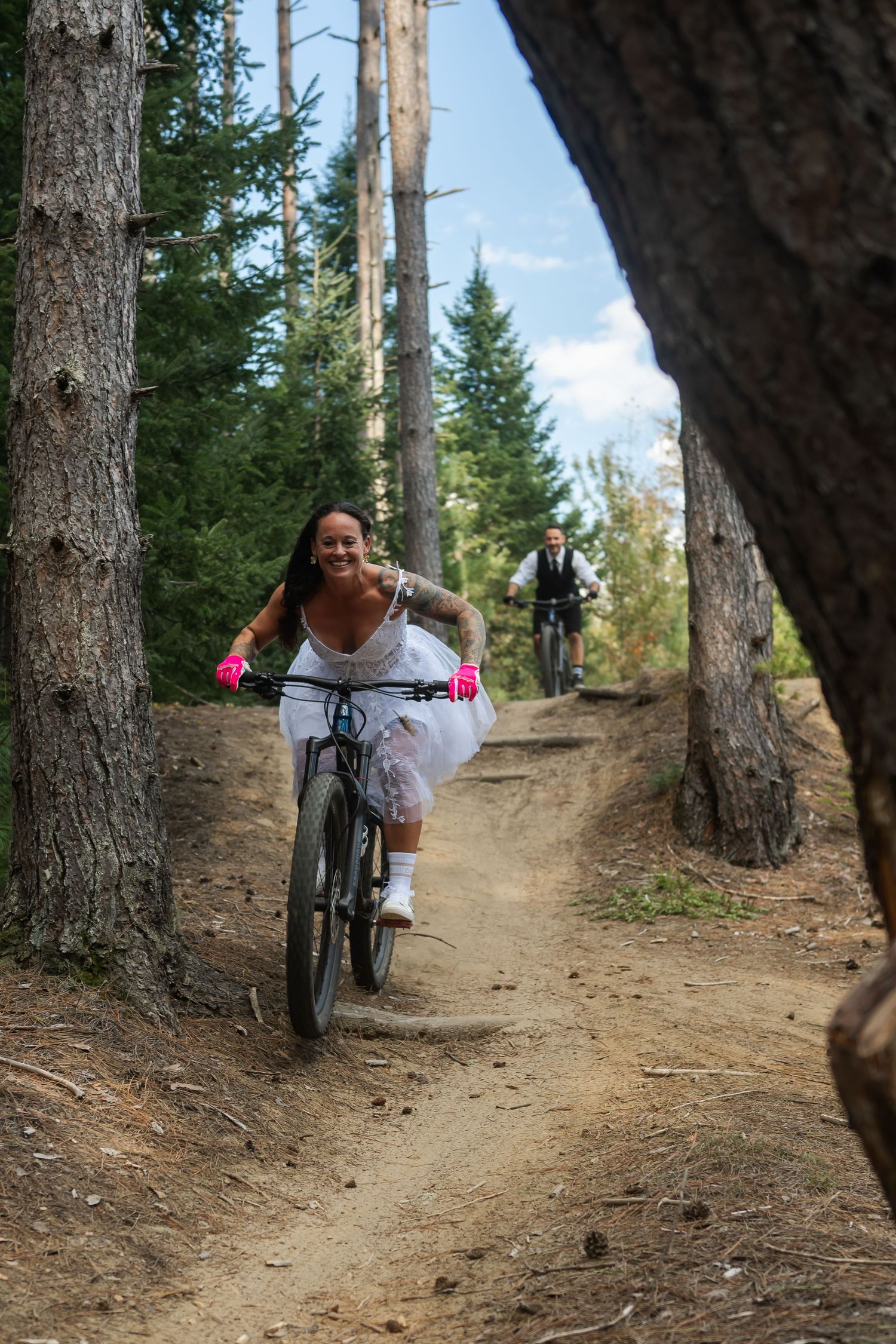 Smiling bride in white dress and pink gloves rides mountain bike down forest trail with groom following behind.