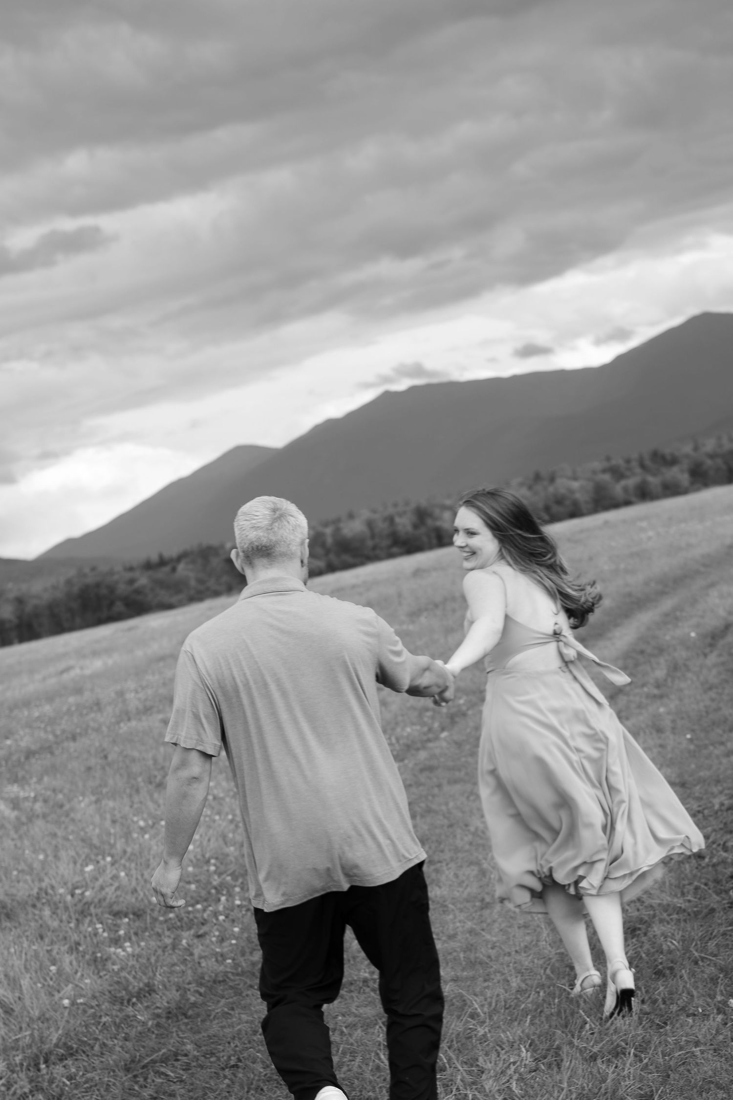 A black and white photo of a couple running through a field, woman in flowing dress looks back smiling.