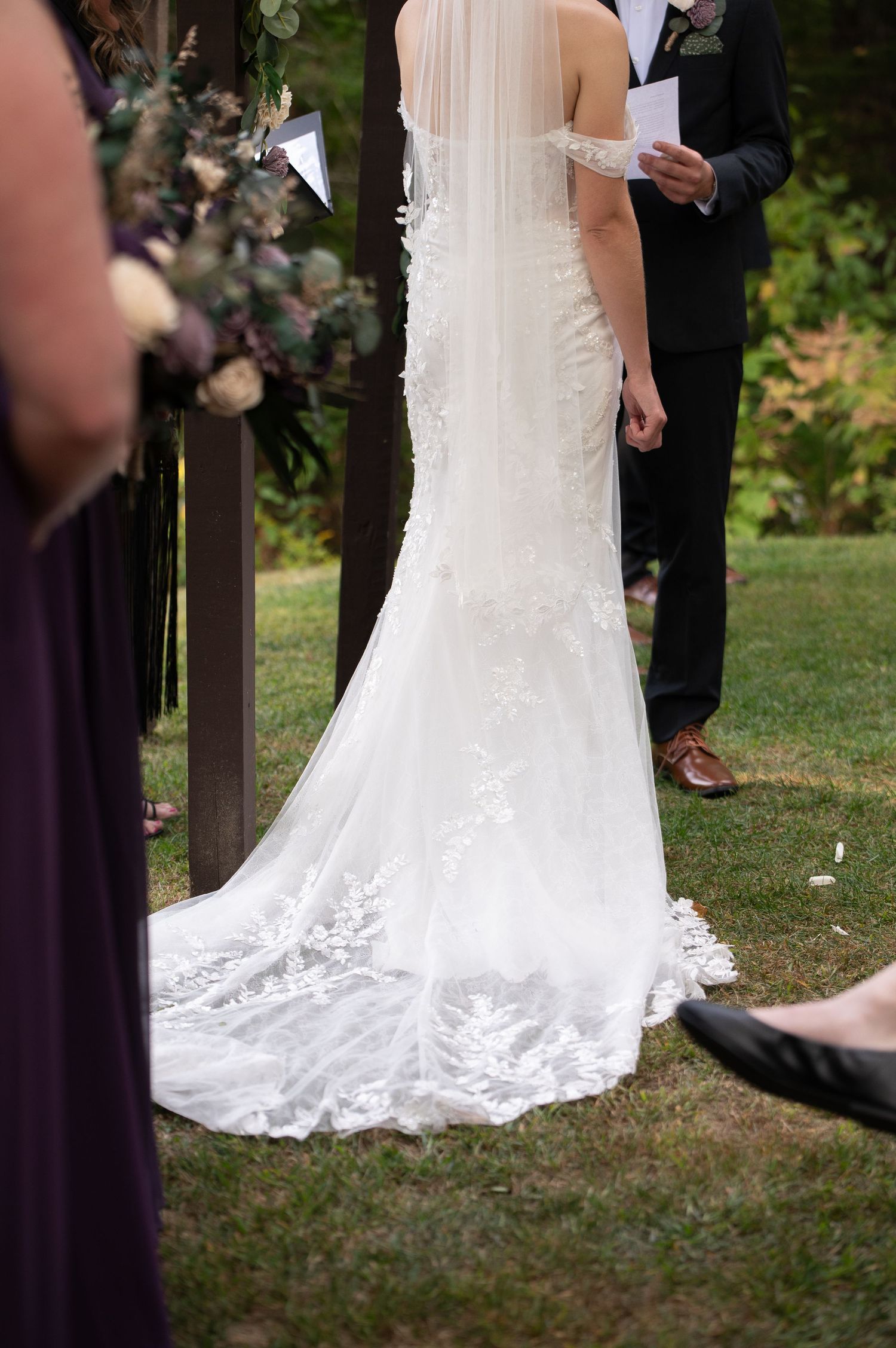Back view of bride in lace wedding gown with train at outdoor ceremony, officiant holding vows nearby.